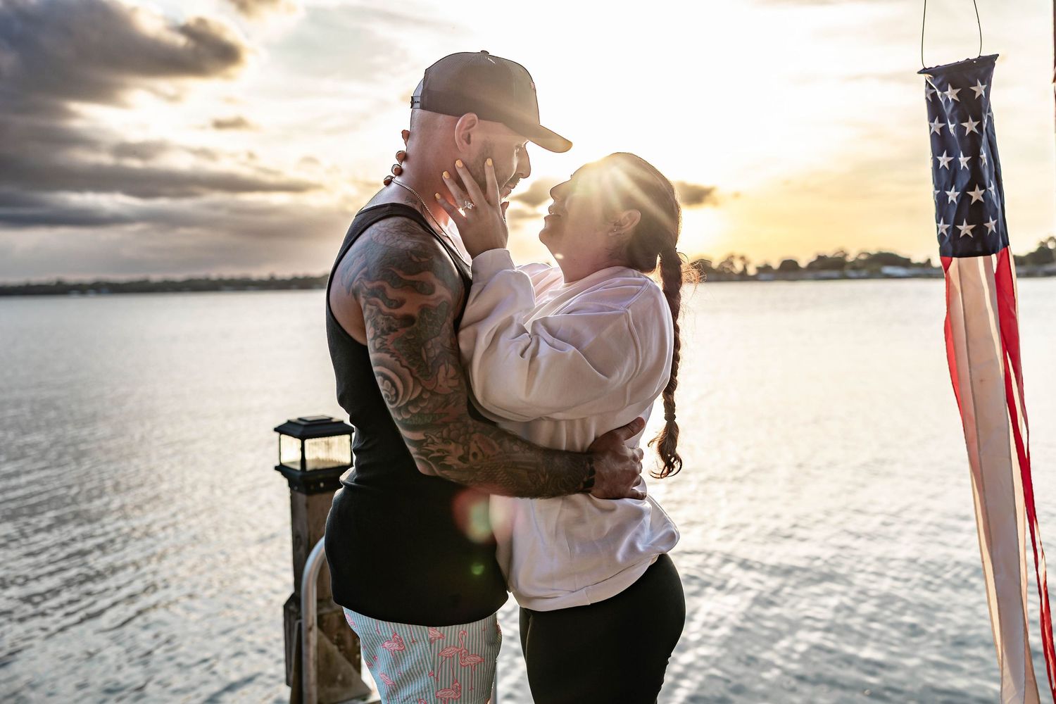 Couple embracing romantically on a waterfront dock at sunset with an American flag in the background.