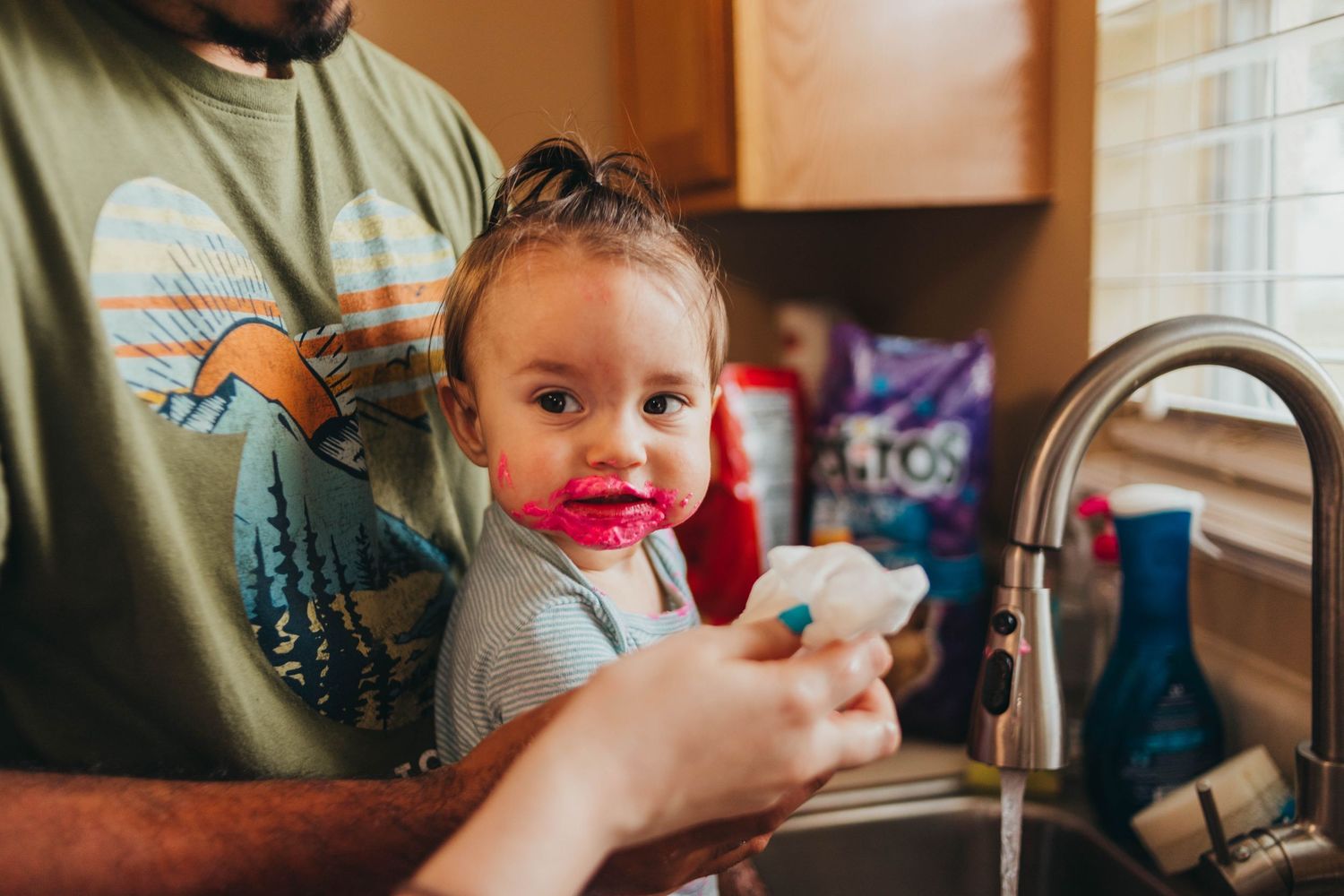 Parent helps young toddler wash hands at kitchen sink during a candid lifestyle family photo.