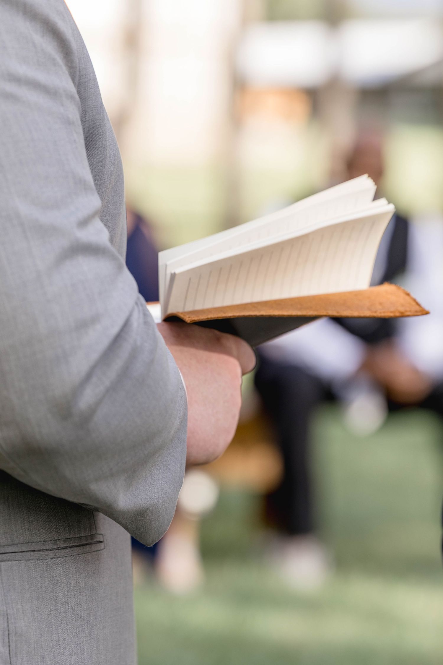 Groom in gray suit reads wedding vows from a leather-bound book during outdoor ceremony.