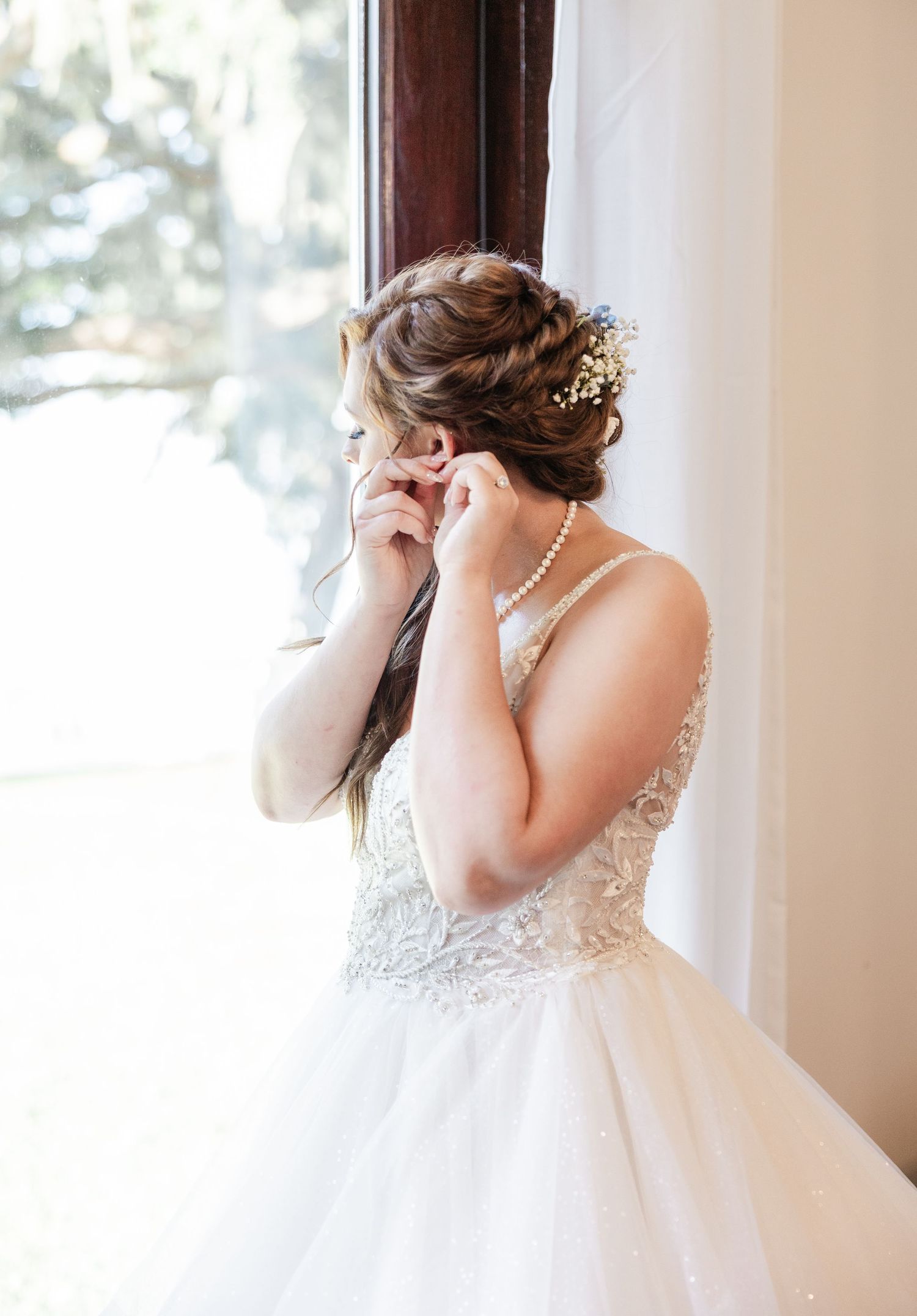 Bride in lace wedding gown adjusting earring by window with elegant updo and pearl necklace.