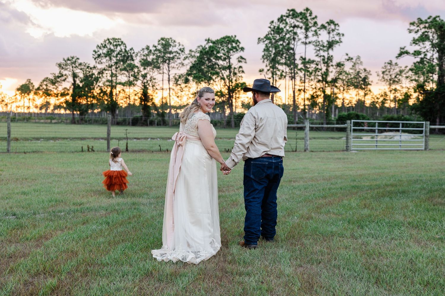 Bride and groom hold hands at sunset on green farm field as toddler in orange plays nearby.