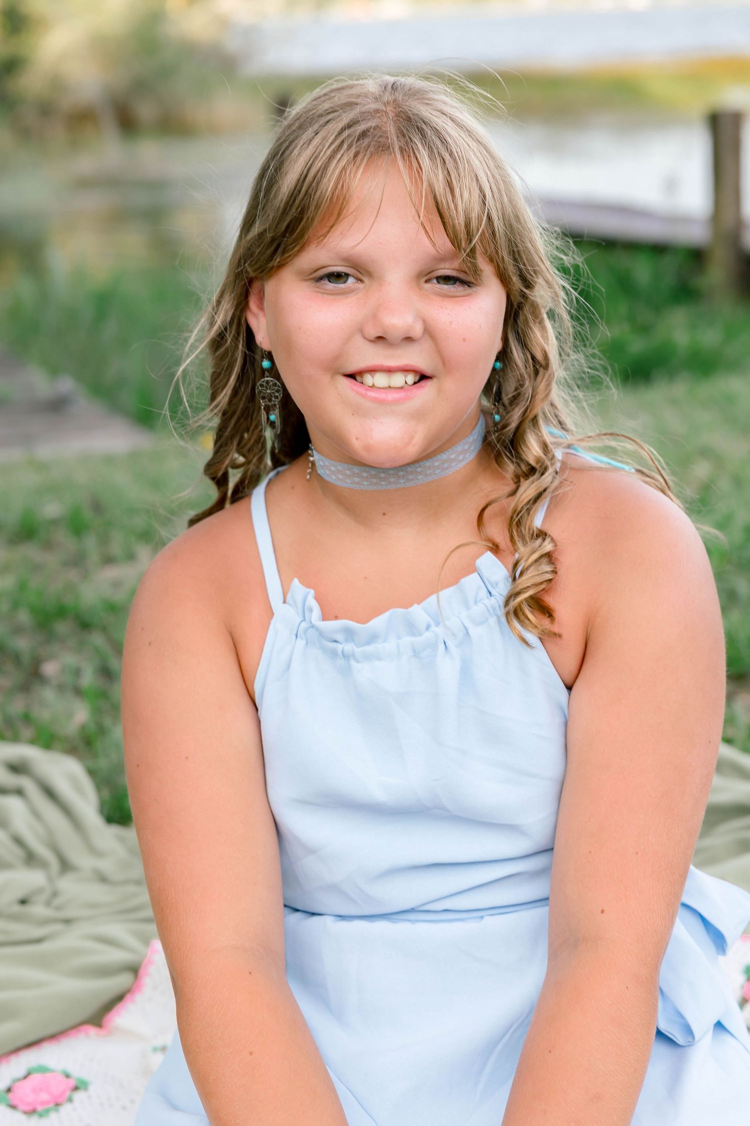 Smiling young girl in light blue dress with turquoise earrings and choker necklace sitting outdoors near water.