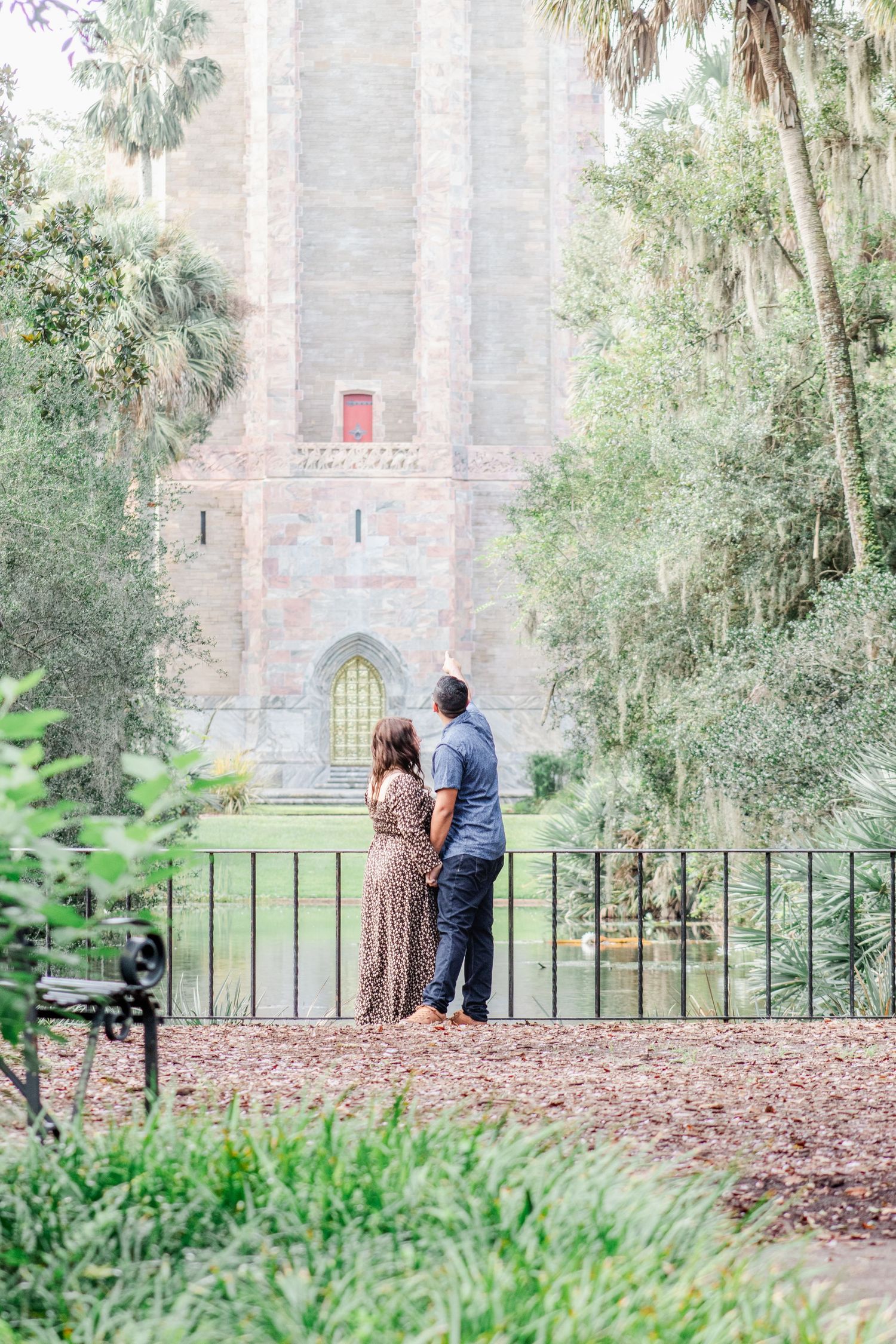 Couple stands together viewing historic stone tower surrounded by lush greenery and palm trees.