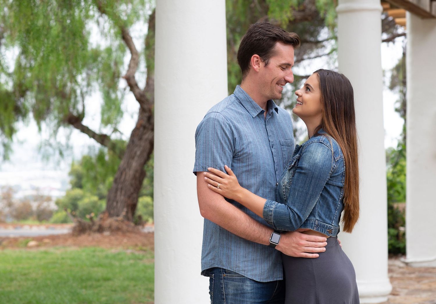 A smiling couple embraces near white columns outdoors, gazing lovingly at each other in warm light.