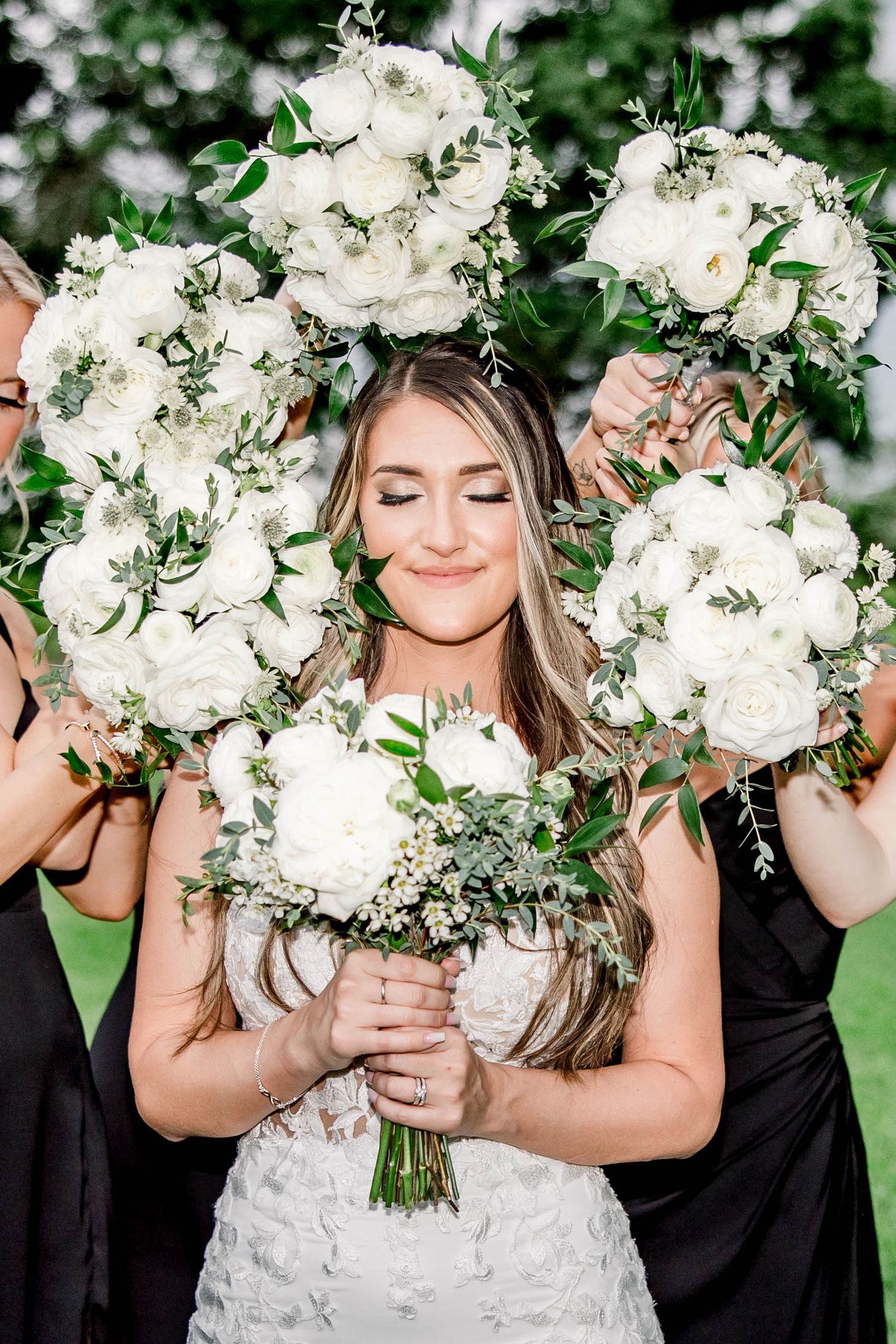 Smiling bride surrounded by bridesmaids holding large white peony and eucalyptus bouquets outdoors.