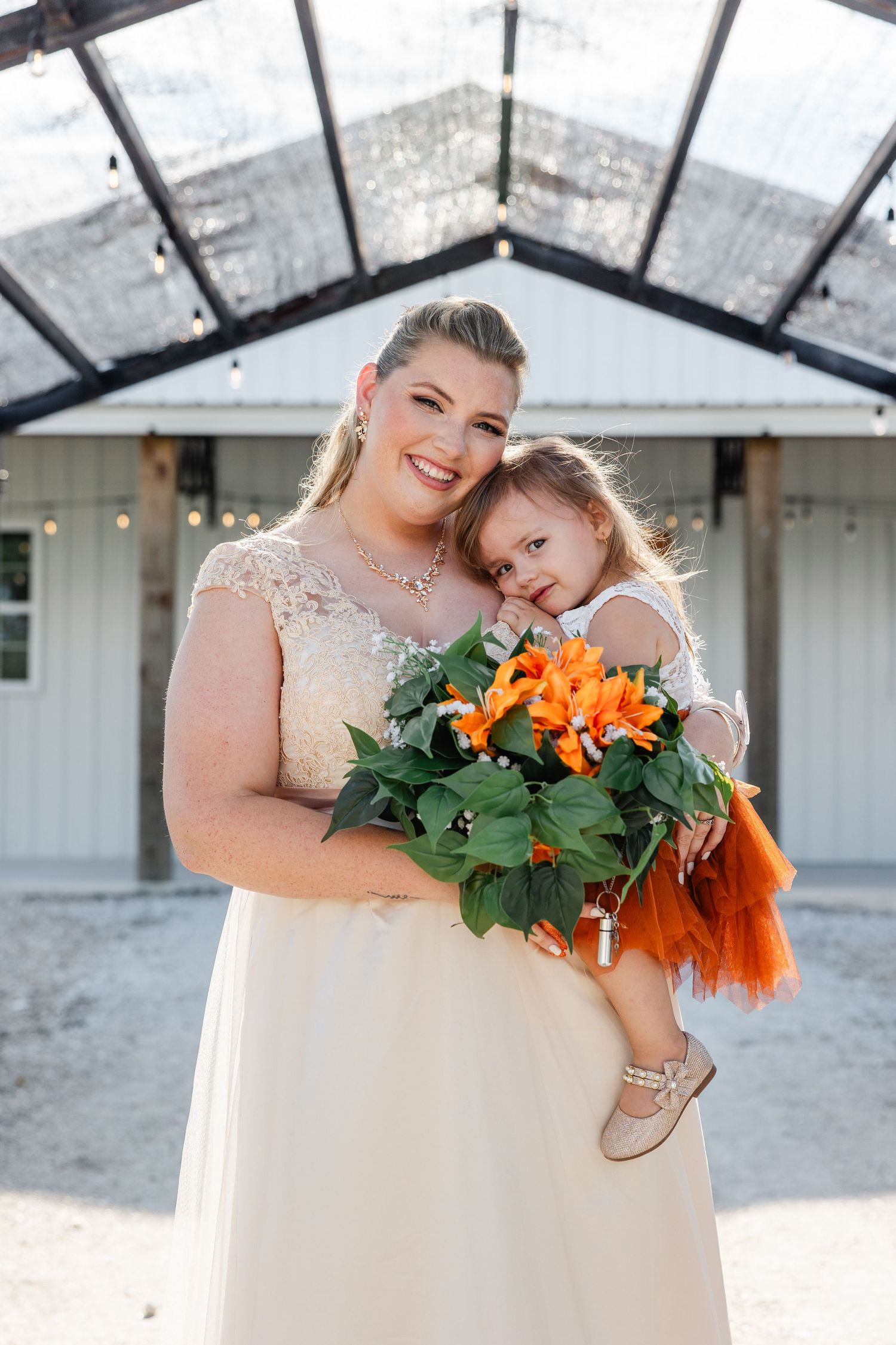 Bride in lace gown holds orange flower bouquet and young girl in orange tutu outside barn venue.