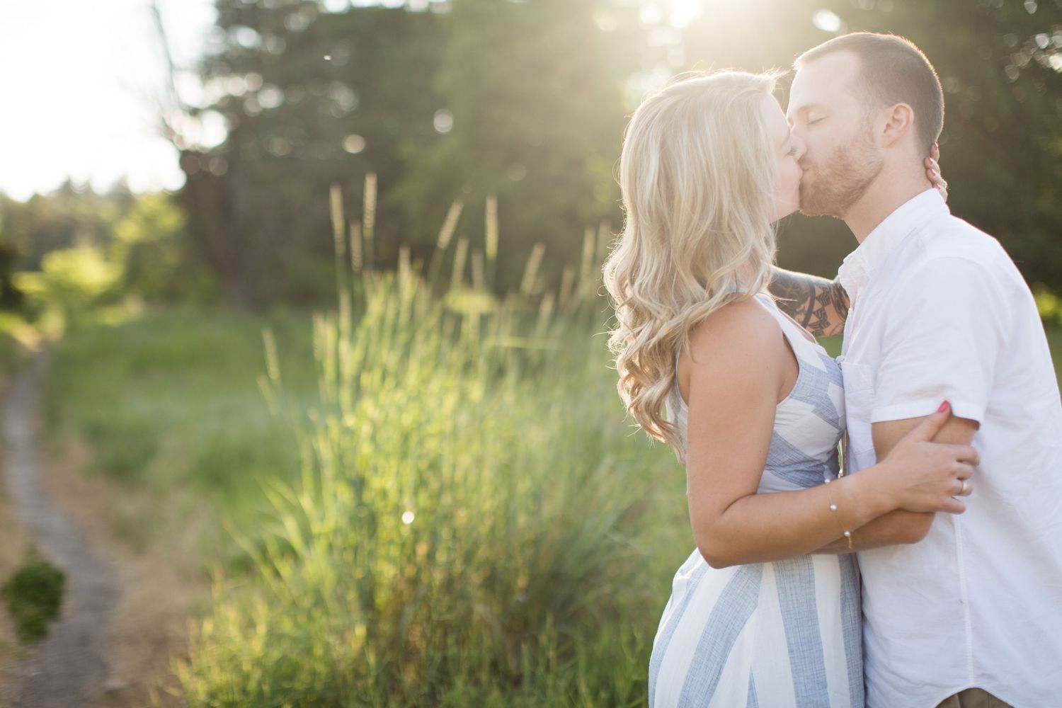A couple shares a romantic kiss in a sunlit meadow, the woman in a light blue dress embracing her partner.