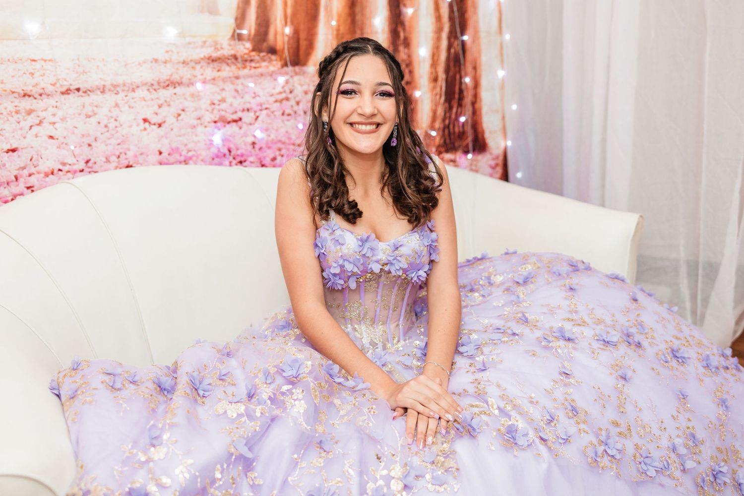Smiling teenage girl in a lavender floral quinceañera gown seated on a white couch with a floral backdrop.