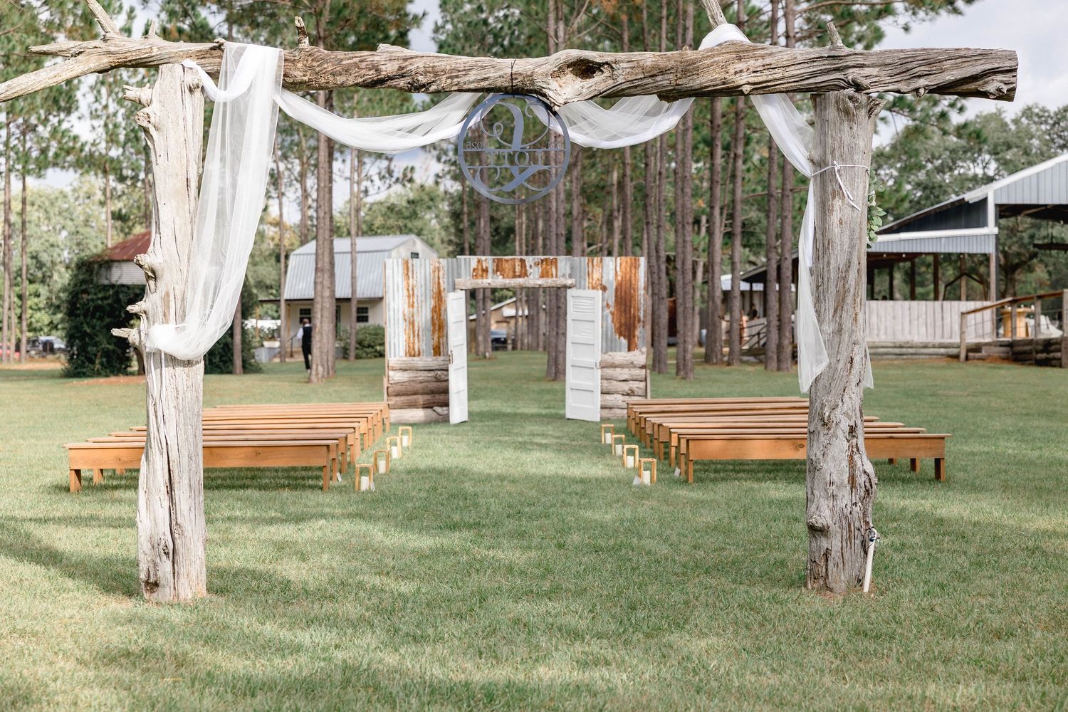 Rustic outdoor wedding ceremony setup with wooden arbor, draped fabric, and wooden pew benches.
