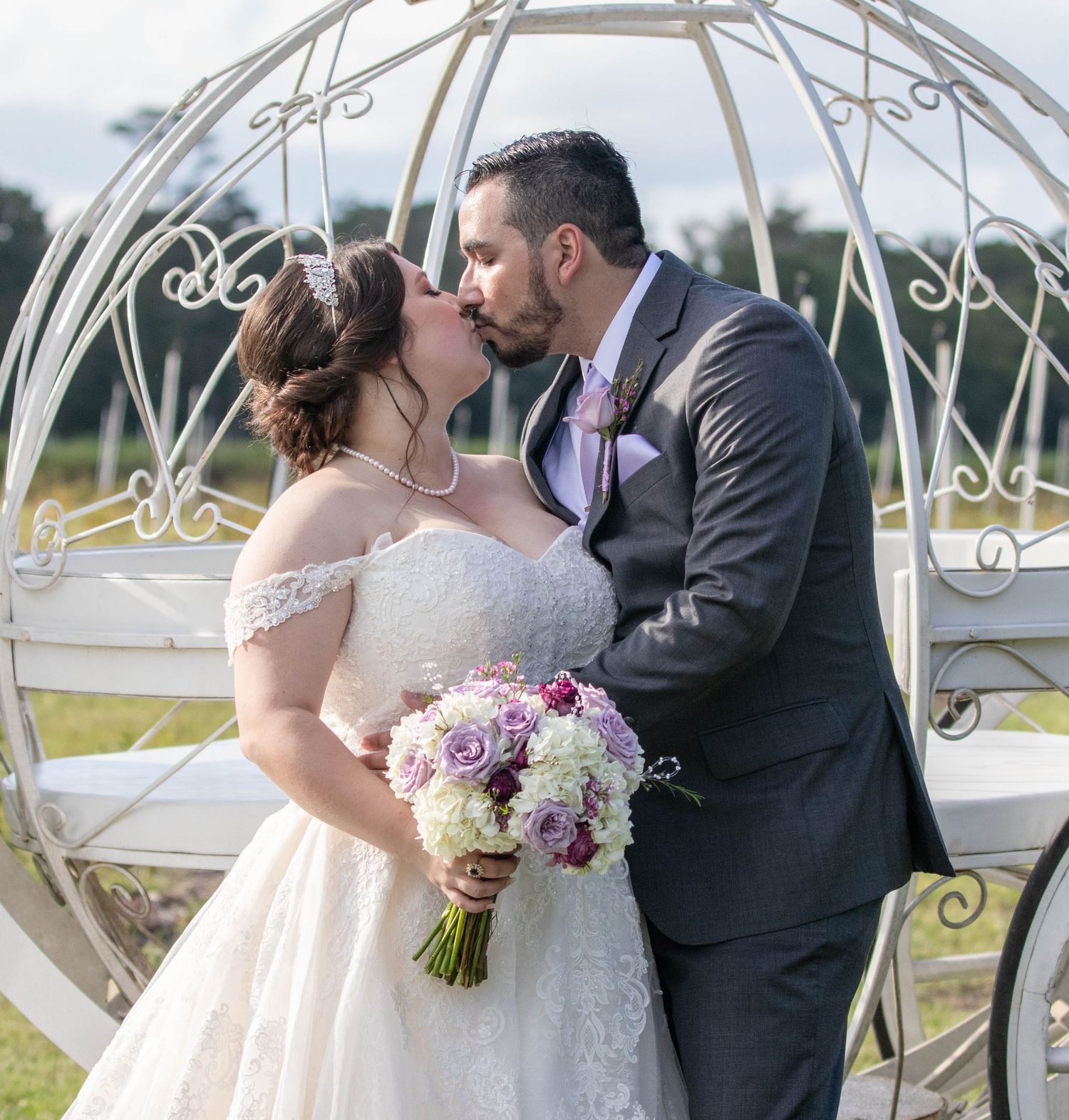 Bride and groom share a kiss beside a white Cinderella carriage with purple rose bouquet.