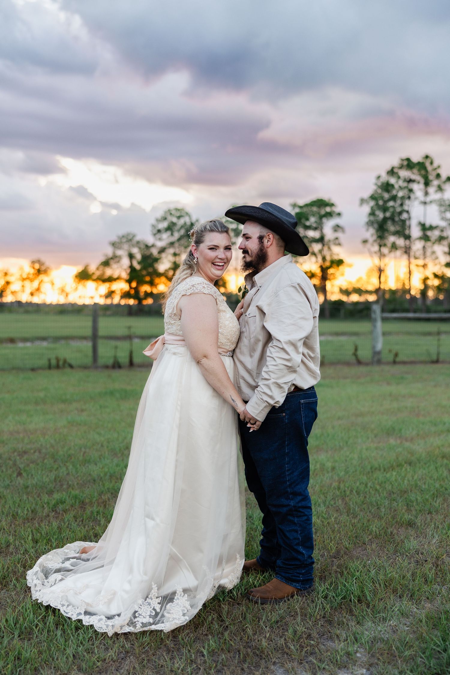 Bride and groom hold hands at golden sunset on a green farm field, groom in cowboy hat.