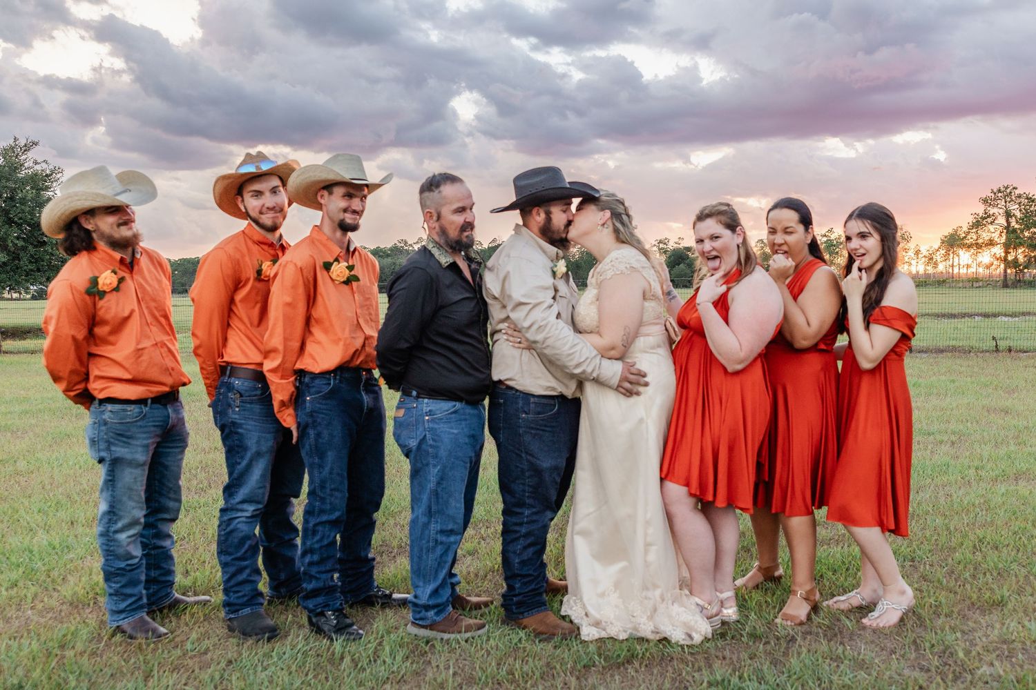 Wedding party in orange and red dresses with cowboy hats celebrate with couple at sunset.