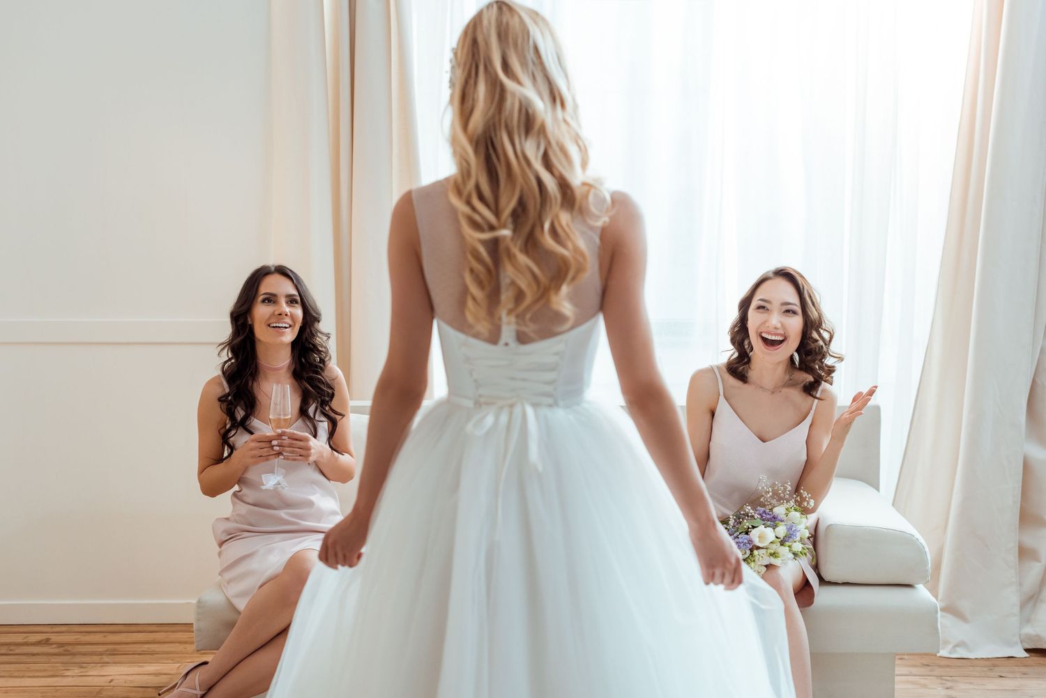 Bridesmaids reacting with joy as bride reveals her white wedding gown in bright elegant room.