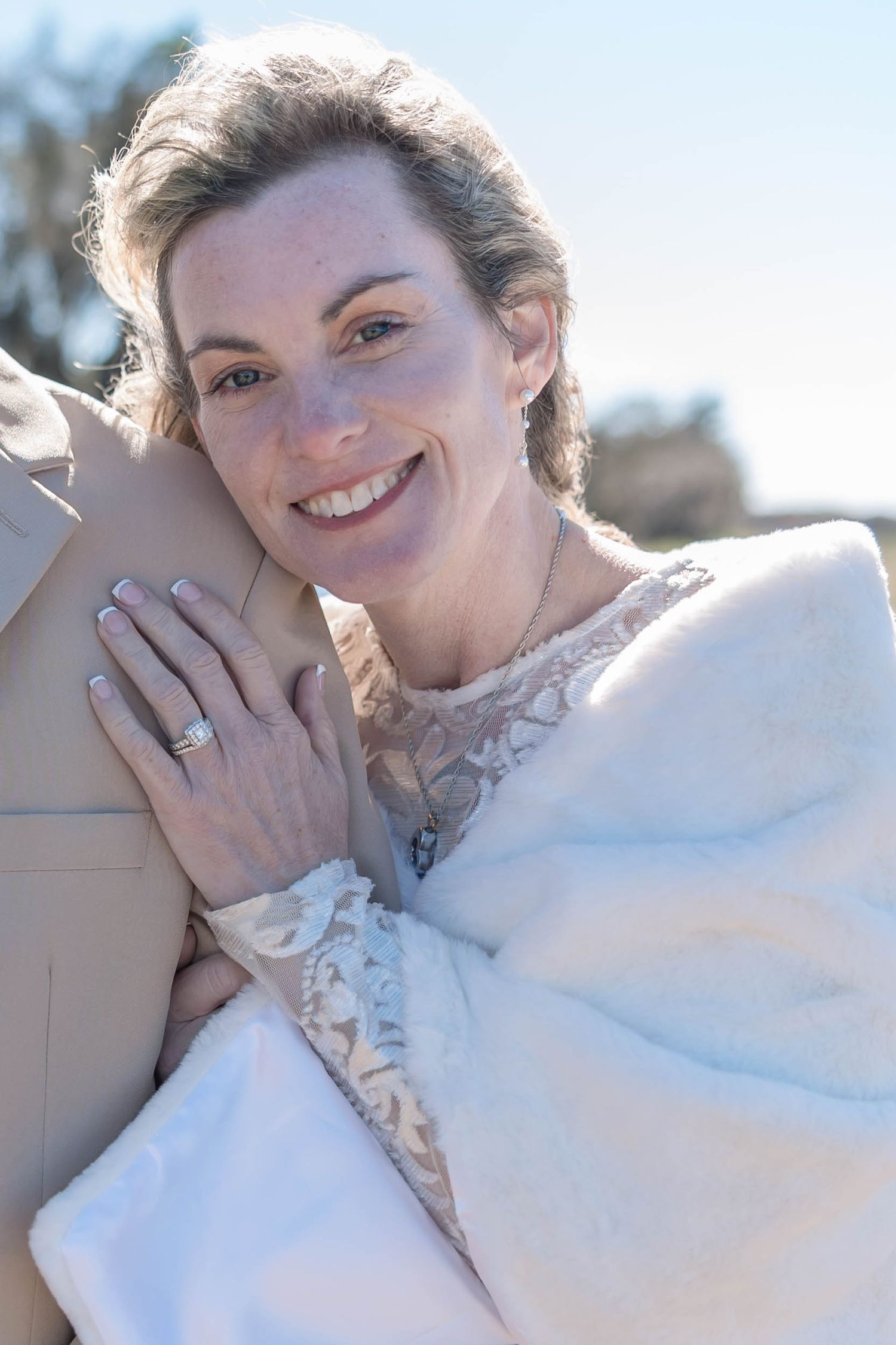 Smiling bride in lace dress and white fur wrap hugging her partner outdoors.