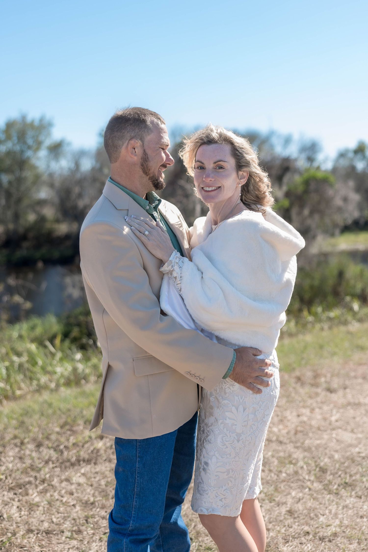 Couple embracing in a sunny outdoor field, woman in white lace dress and fur wrap.