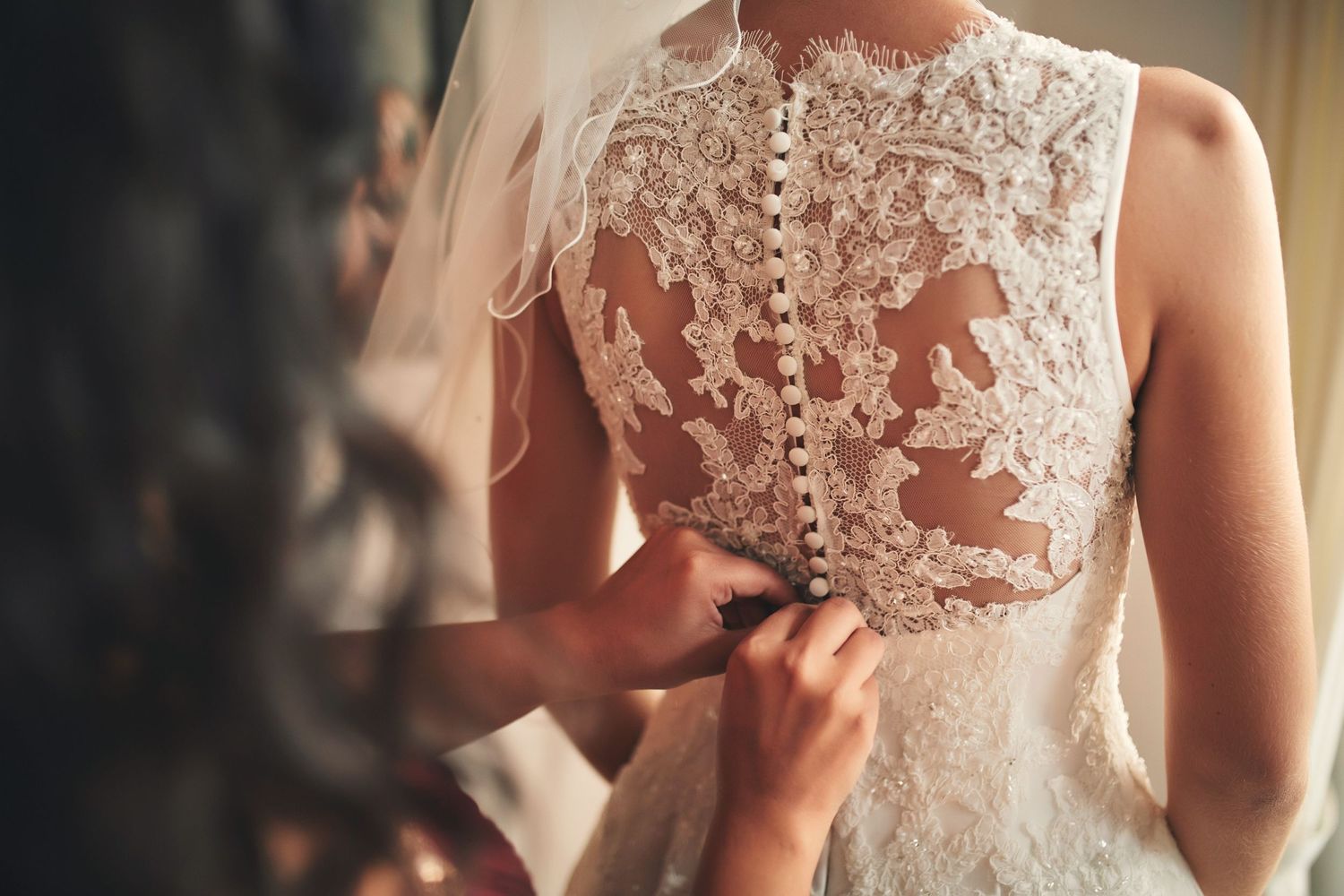 Close-up of intricate lace wedding dress being buttoned up the back by a bridesmaid.