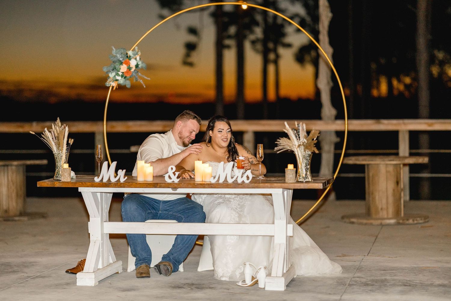 Newlyweds cut wedding cake at sweetheart table with golden hoop backdrop at sunset outdoor reception.