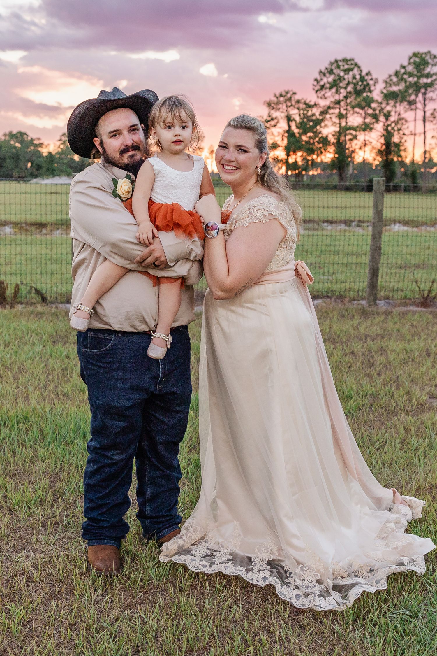 Couple in wedding attire poses with young child at golden hour on a rustic farm setting.