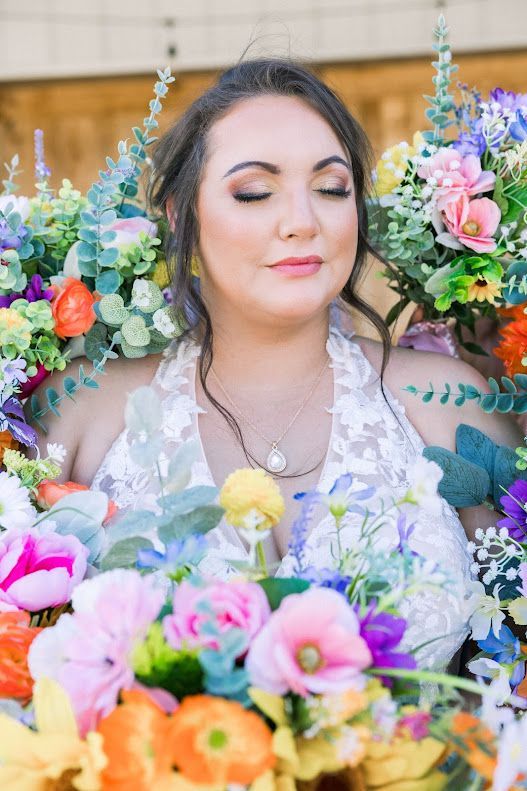 Bride with closed eyes surrounded by colorful wedding bouquet flowers in white lace dress.
