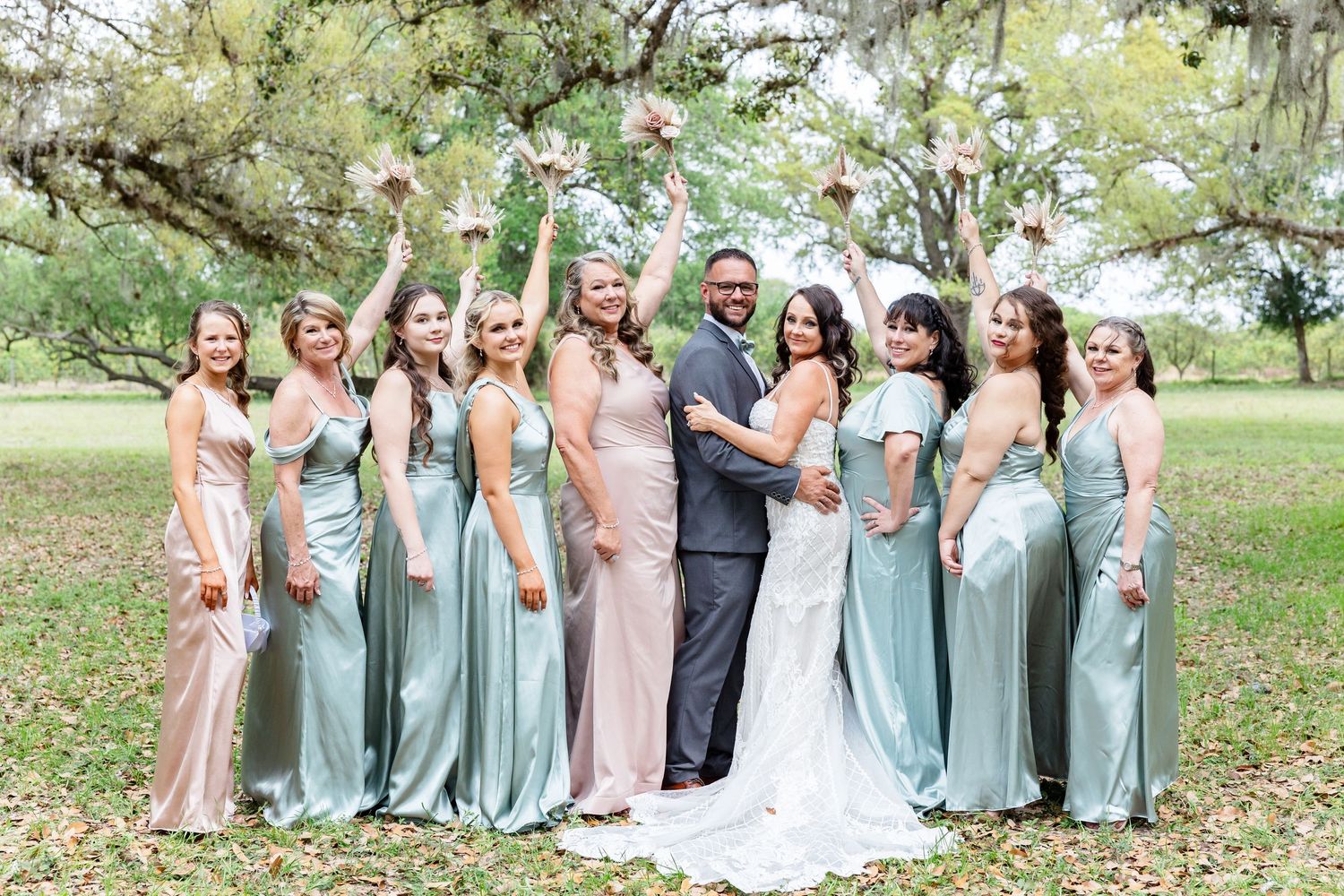 Wedding party in sage green and blush bridesmaid dresses celebrating under large oak trees.