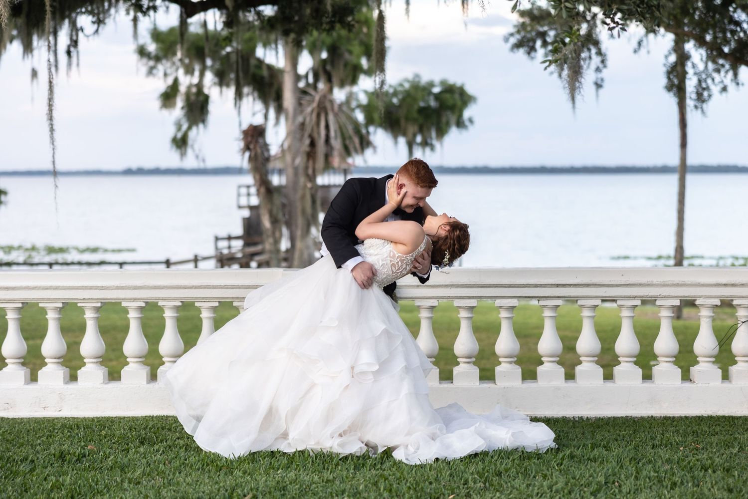 A groom dips his bride in a dramatic kiss with a scenic waterfront and Spanish moss backdrop.