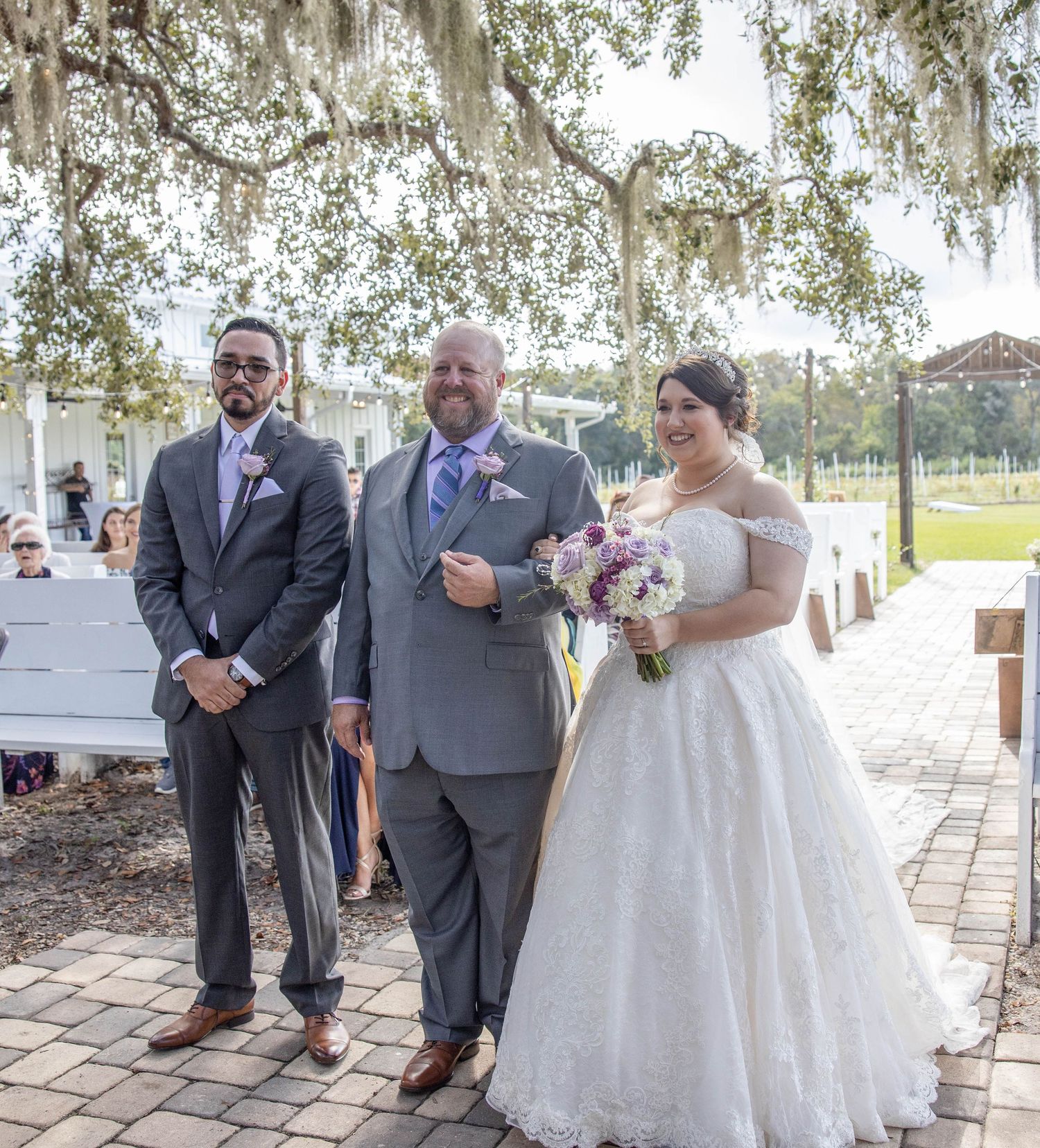 Bride holding purple bouquet walks wedding aisle with two escorts under moss-draped trees at outdoor ceremony.