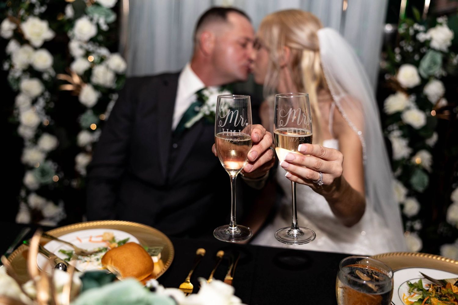 Newlyweds kiss behind Mr. and Mrs. champagne glasses at elegant floral wedding reception.