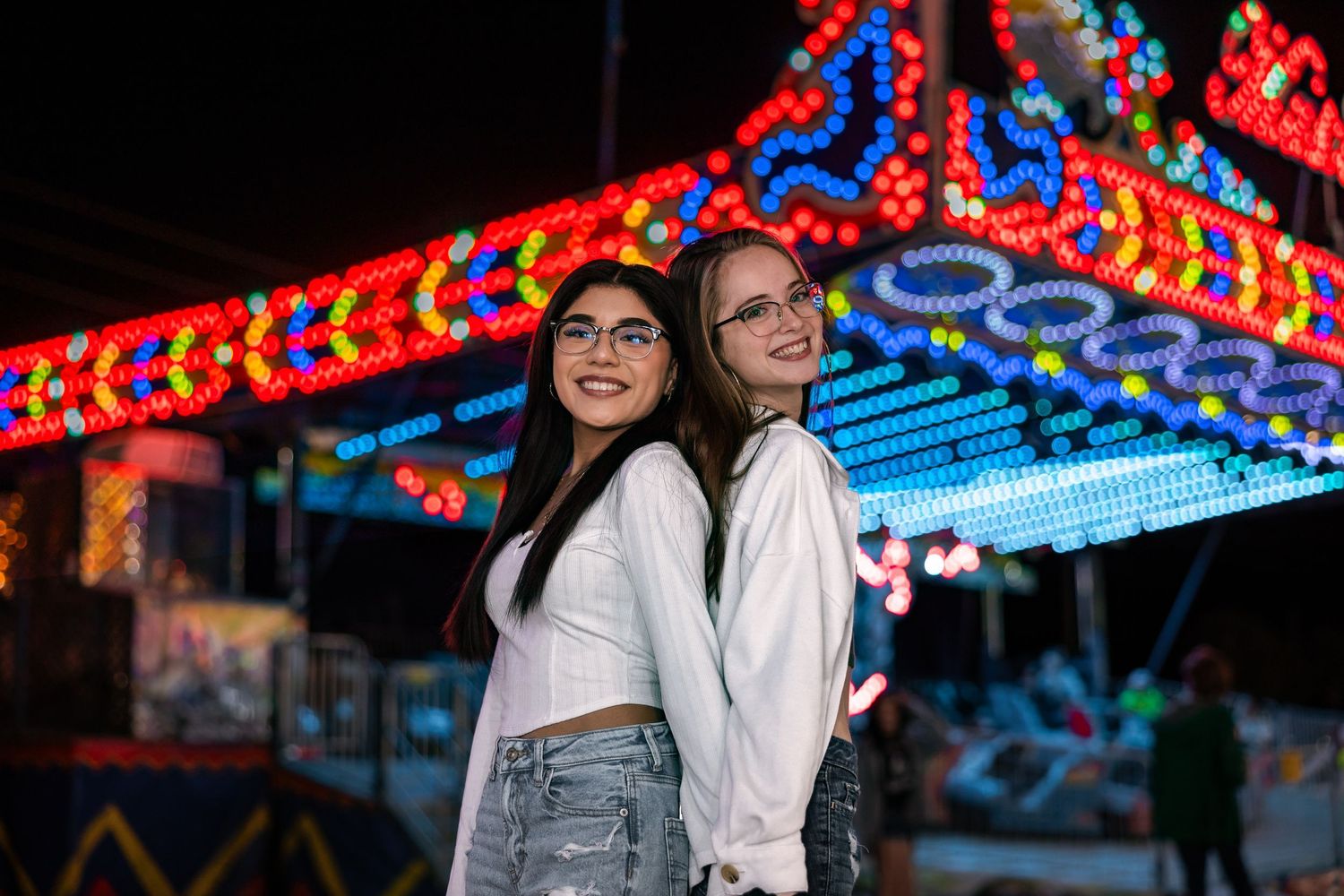Two young women in white shirts smile together in front of colorful carnival ride lights at night.