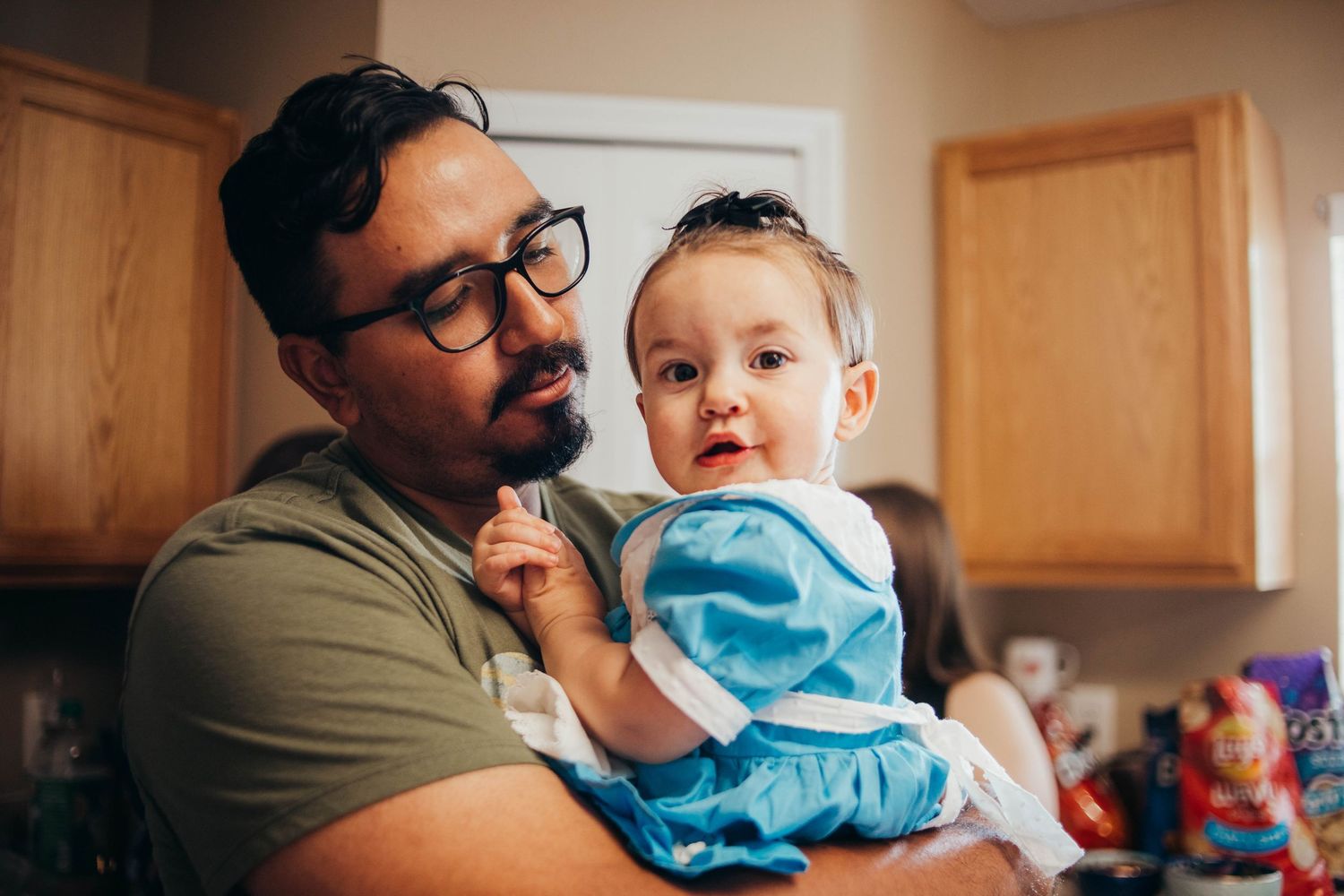 Father wearing glasses holds his baby dressed in a blue outfit in a cozy kitchen setting.