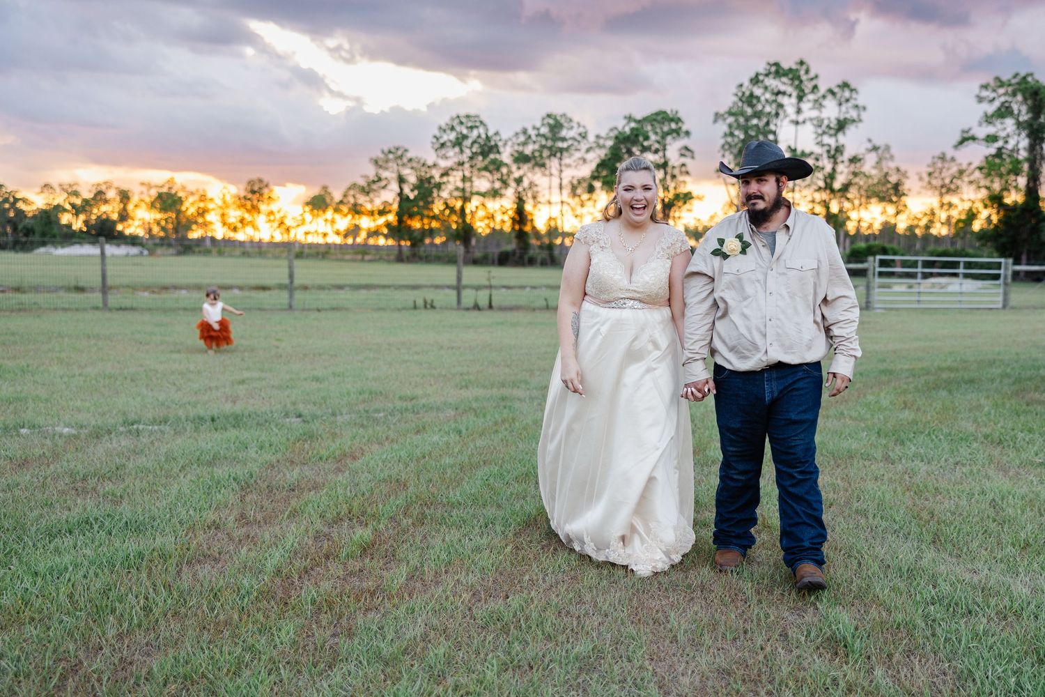 Bride and groom walk hand in hand across green field at golden sunset on rural farm property.
