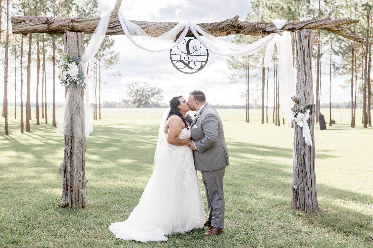 Bride and groom share first kiss under rustic wooden arch with white draping at outdoor wedding ceremony.