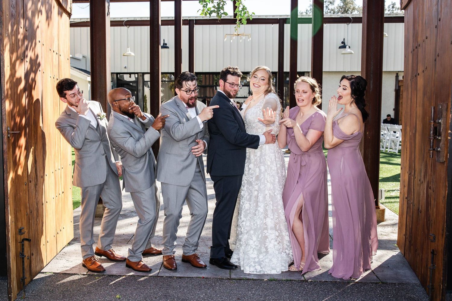 Wedding party in gray suits and mauve dresses celebrating in barn doorway.