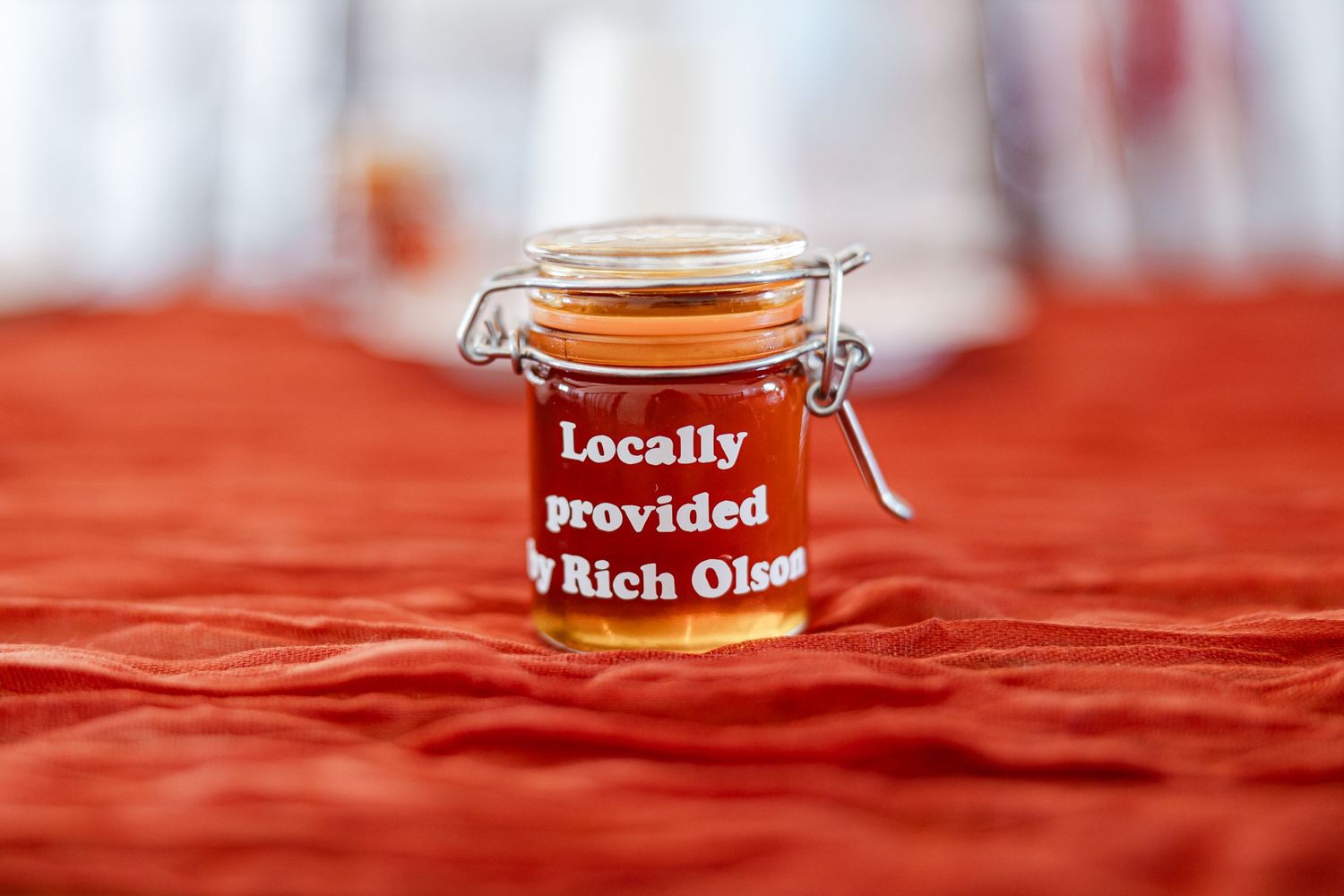 Small glass jar of local honey labeled locally provided by Rich Olson on red tablecloth.