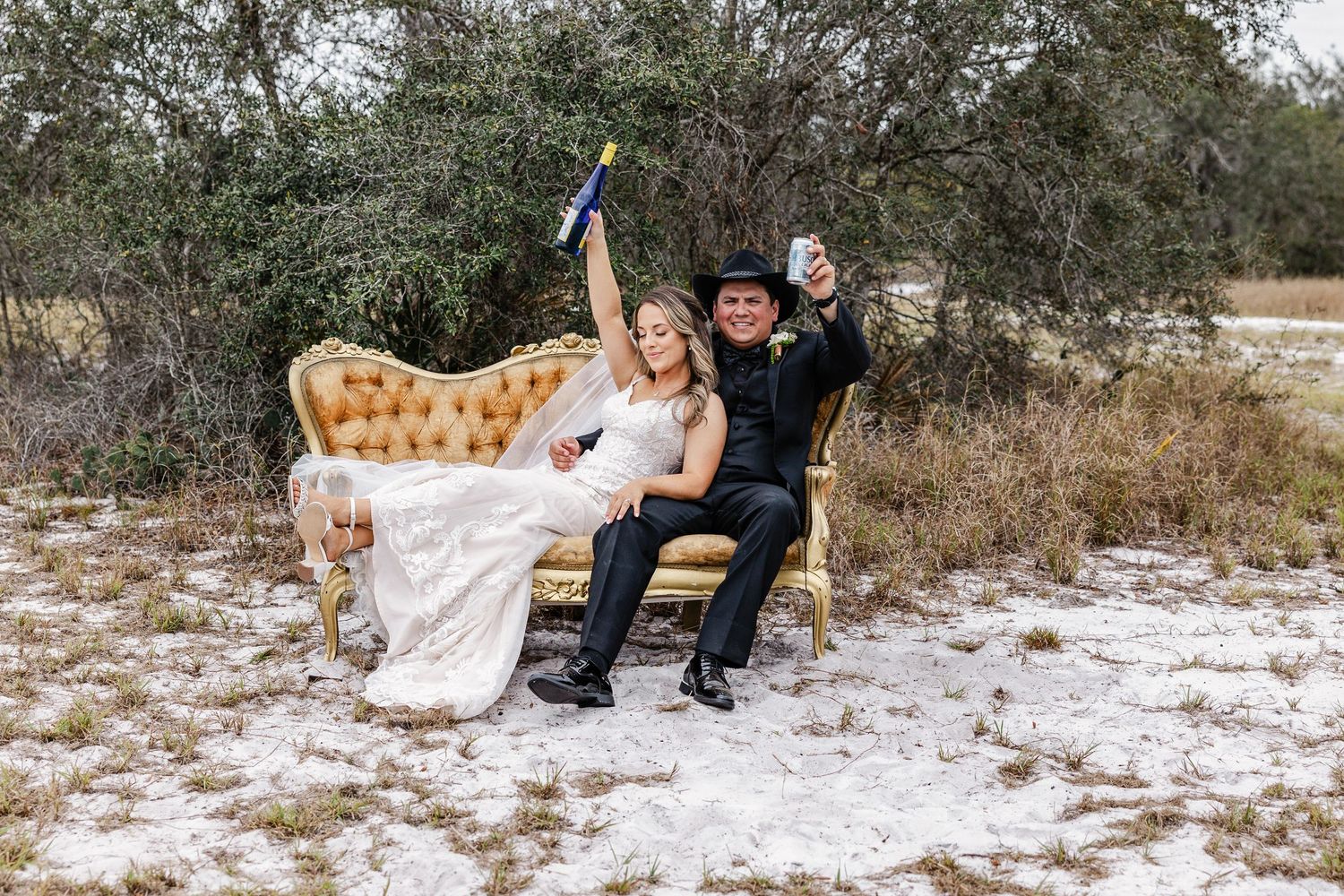 Bride and groom celebrating on a gold vintage couch outdoors, raising drinks with joy on their wedding day.