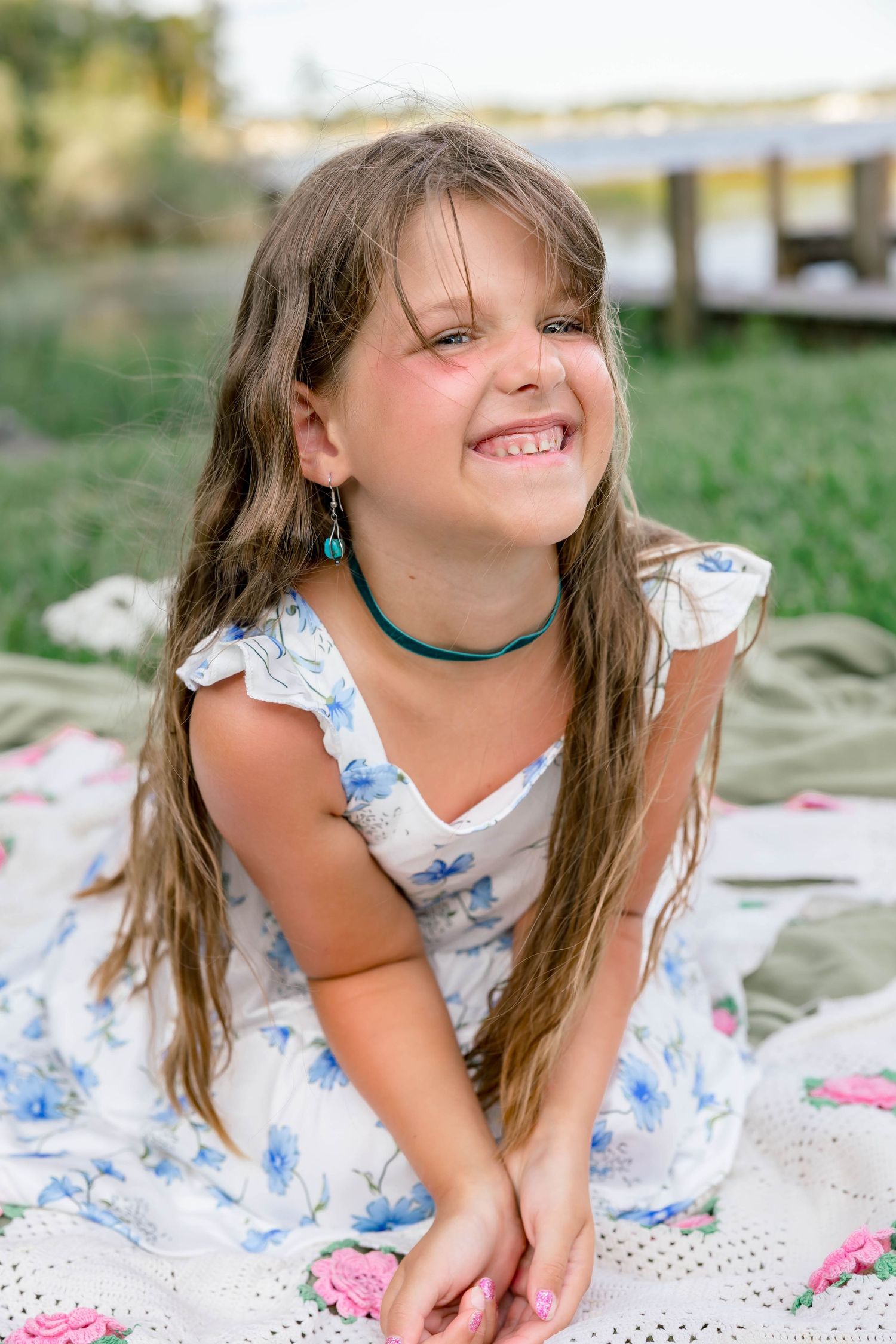 Smiling young girl with long hair wearing a floral dress and turquoise necklace sitting on a blanket outdoors.