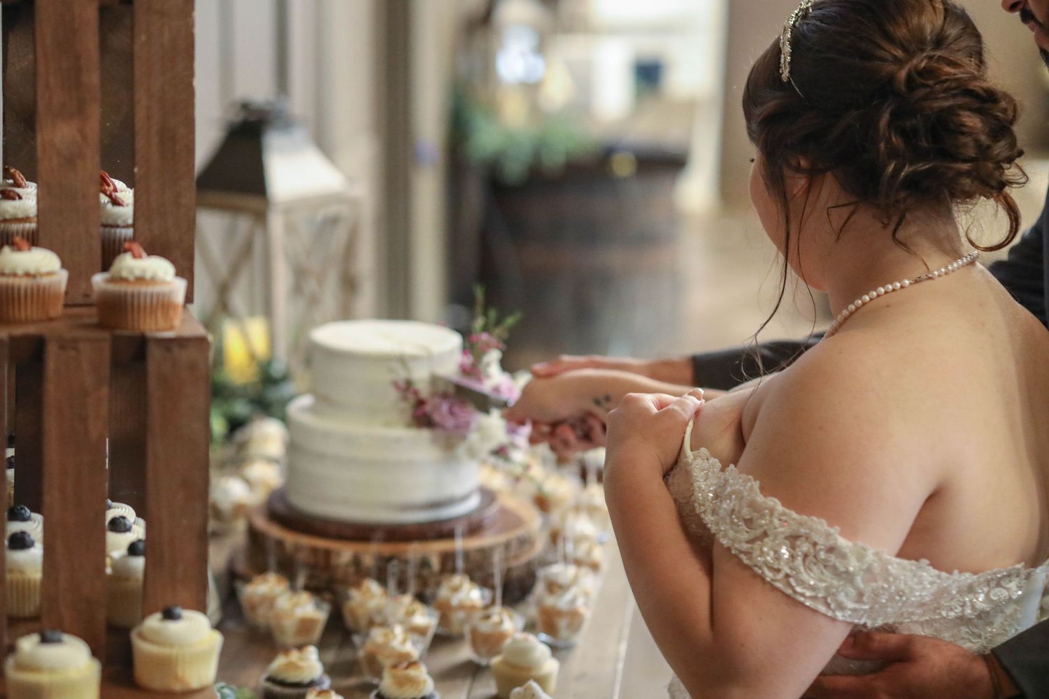 Bride in lace off-shoulder gown cuts wedding cake surrounded by cupcake tower display.
