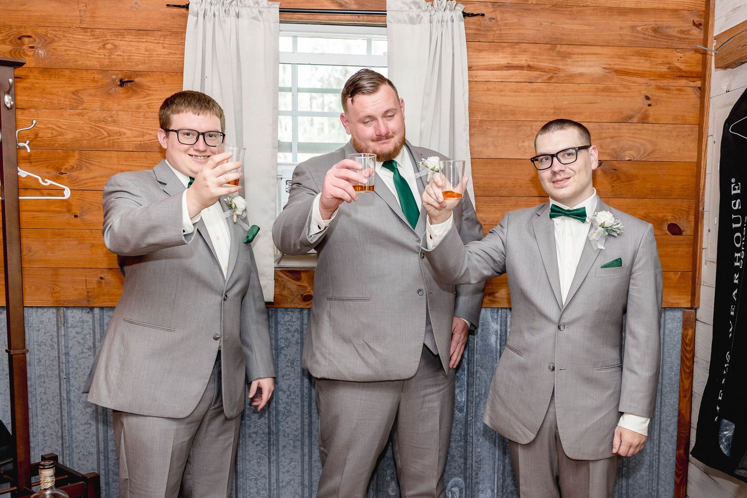 Three groomsmen in gray suits with green bow ties toasting with whiskey glasses before a wedding.