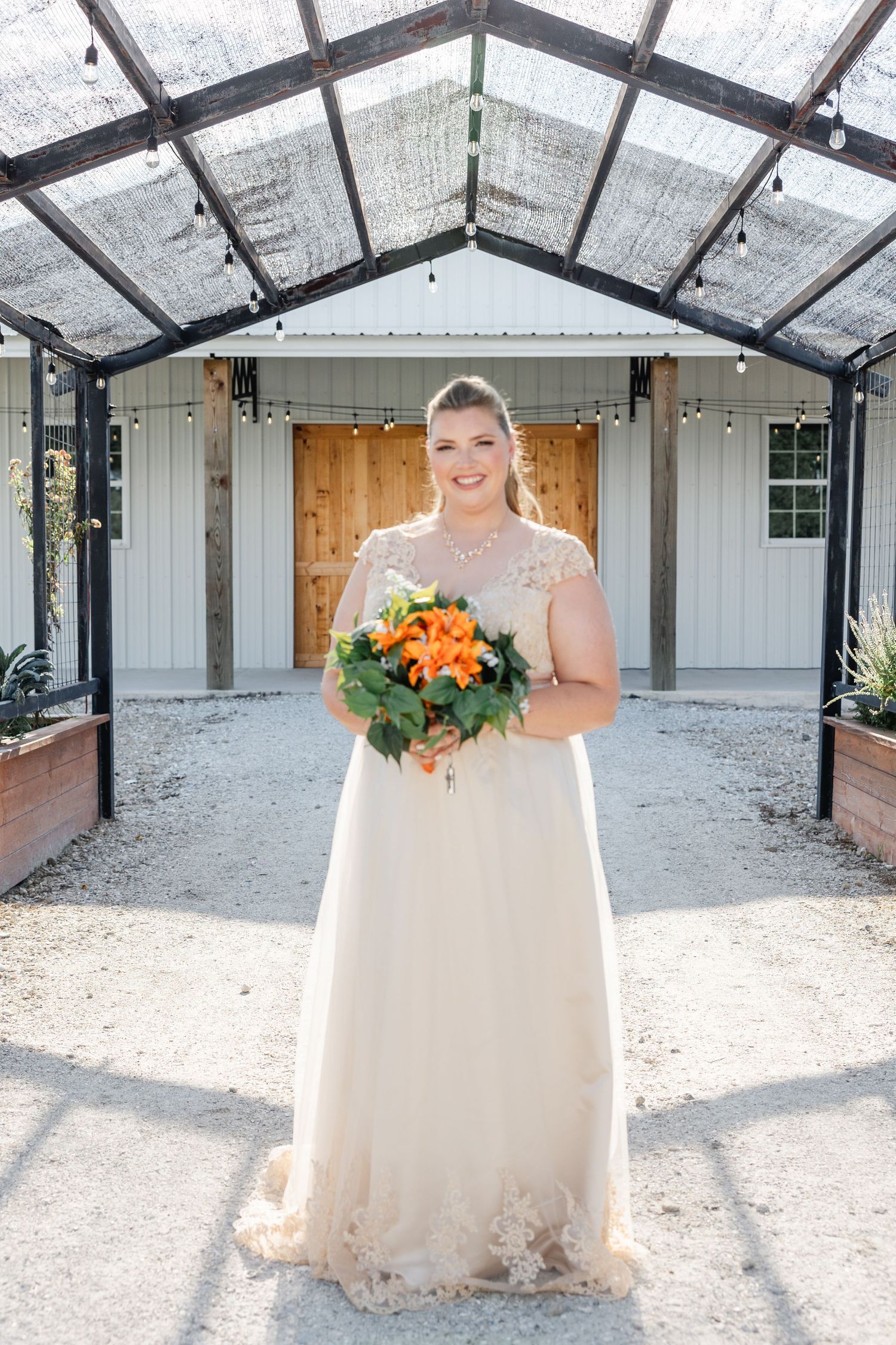 Smiling bride in lace wedding gown holding orange floral bouquet in greenhouse venue.