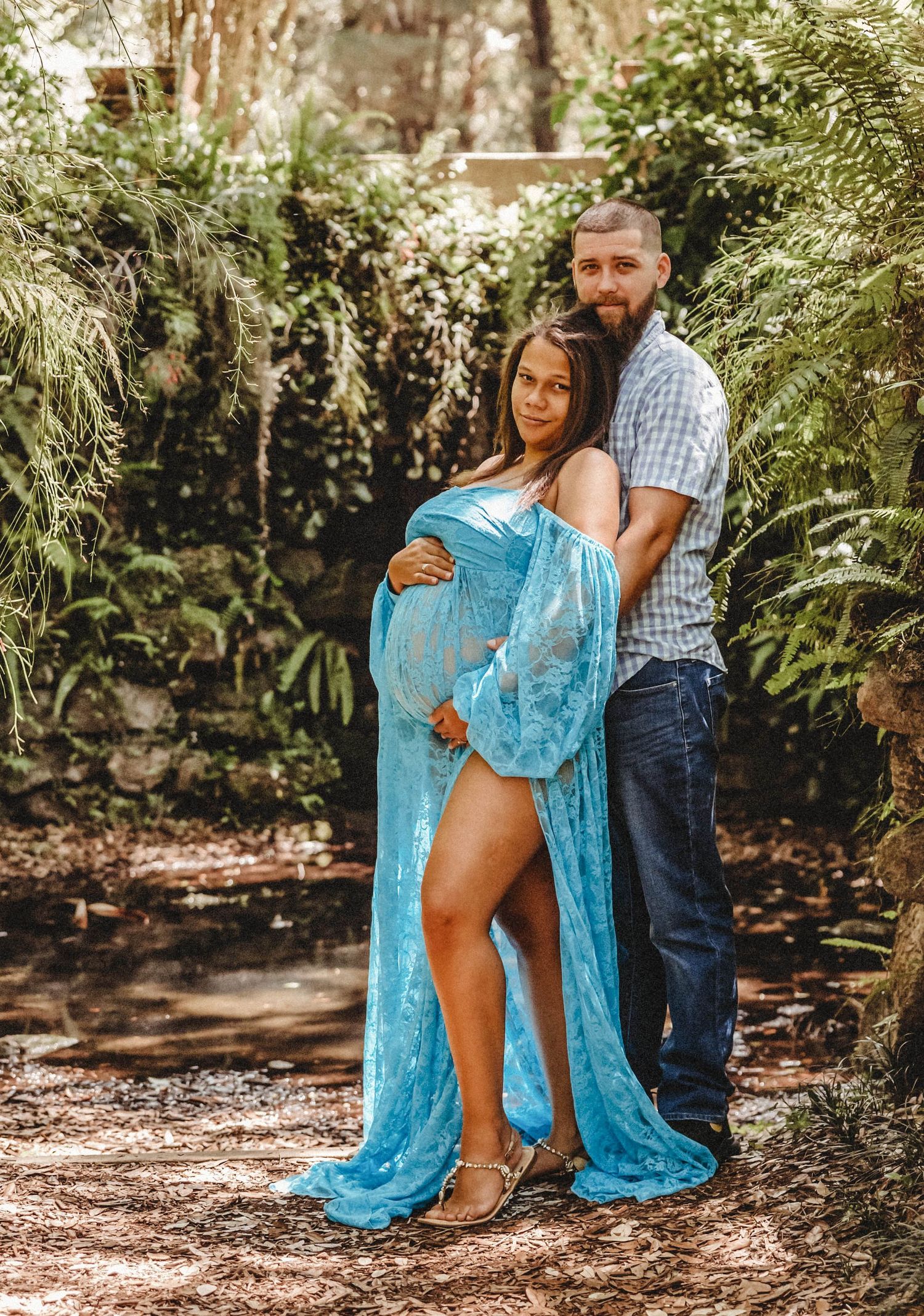 Pregnant woman in blue lace maternity gown posing with partner in a garden.