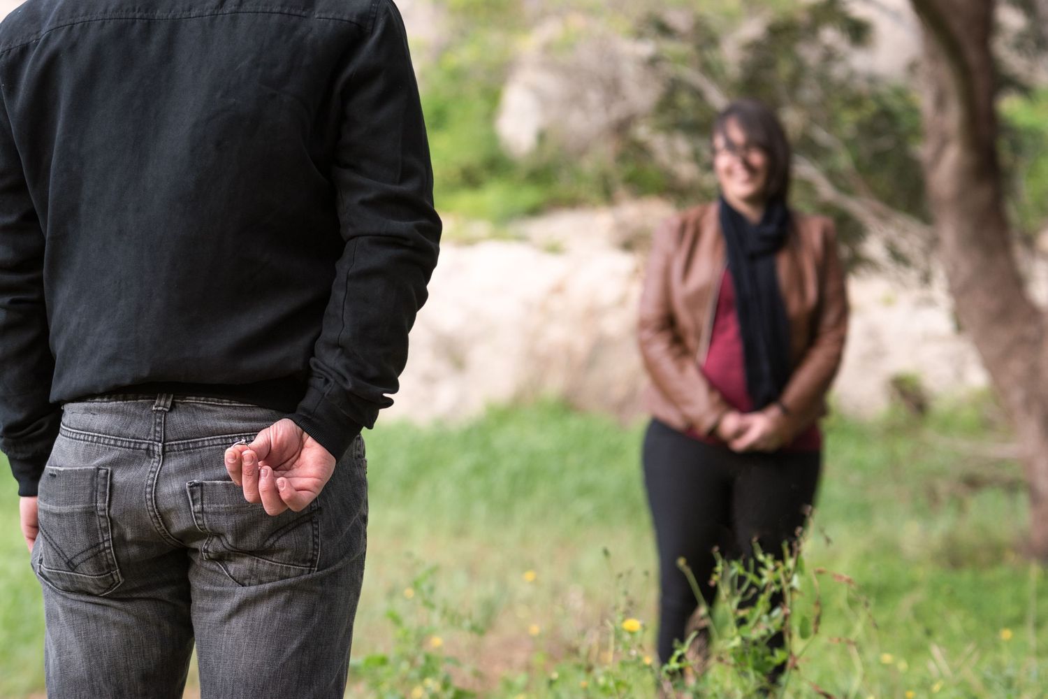 A man hides something behind his back while facing a woman standing in a natural outdoor setting.