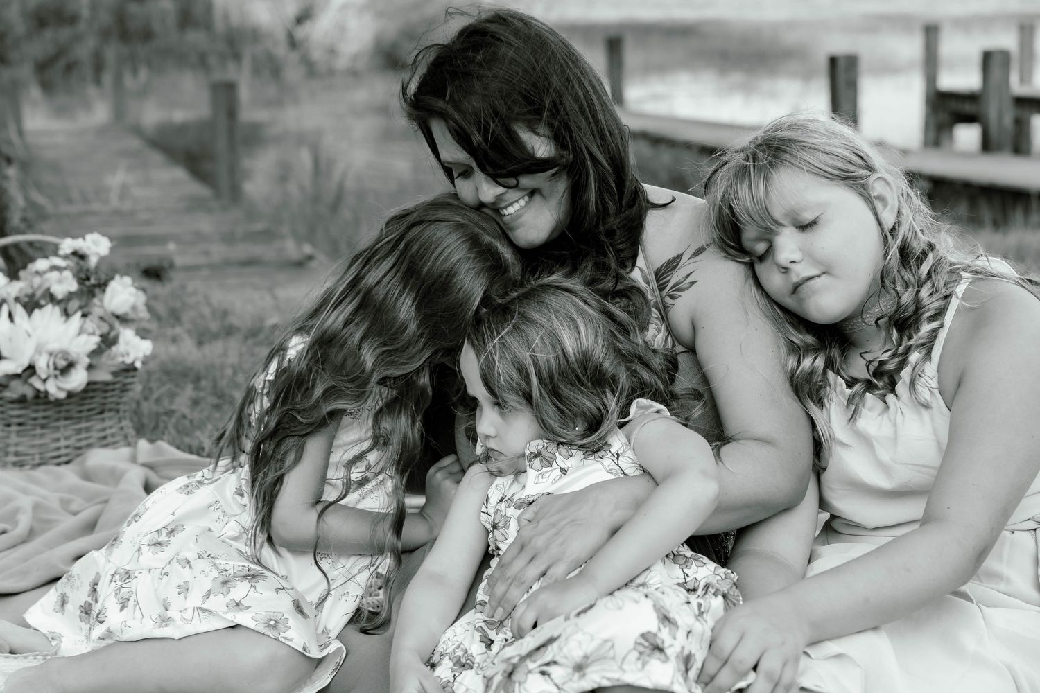 Black and white photo of a mother tenderly embracing her three daughters outdoors near a wooden dock.