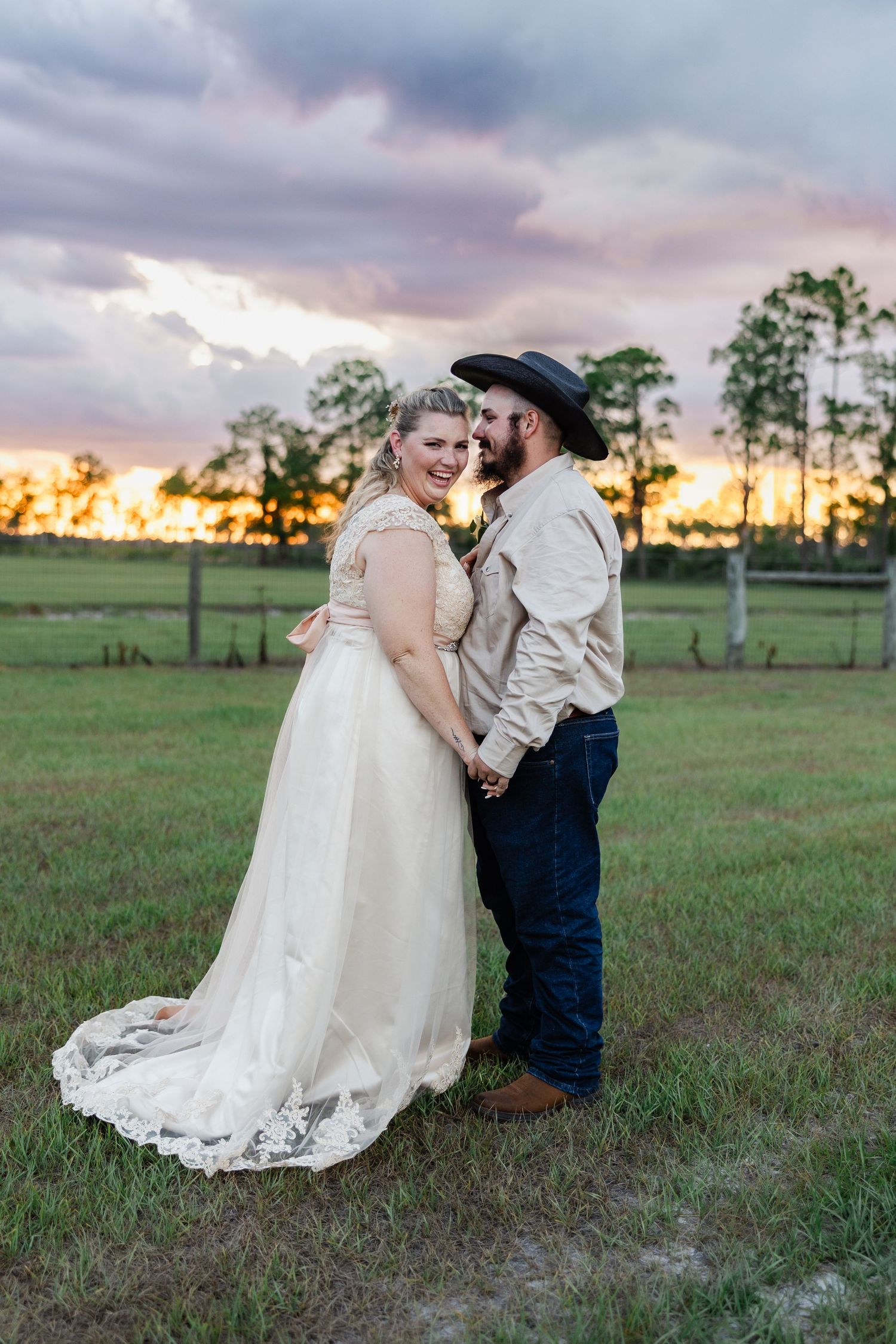 Groom in cowboy hat and bride in lace gown pose holding hands at vibrant pink and orange sunset.