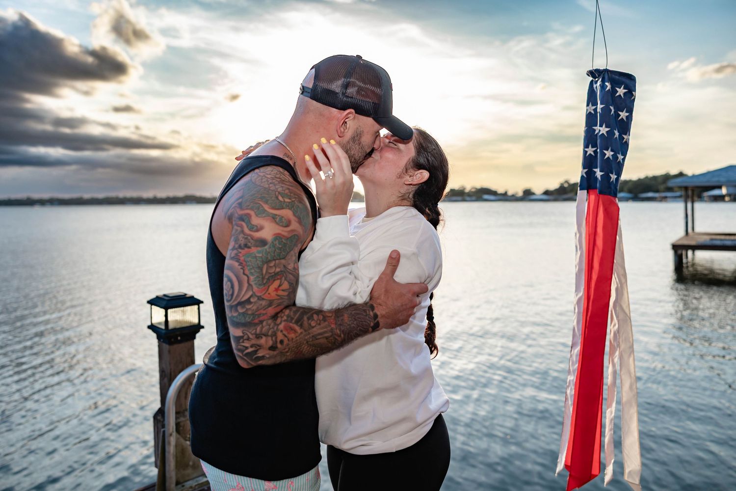 A tattooed man and woman share a passionate kiss on a waterfront dock at sunset near an American flag.