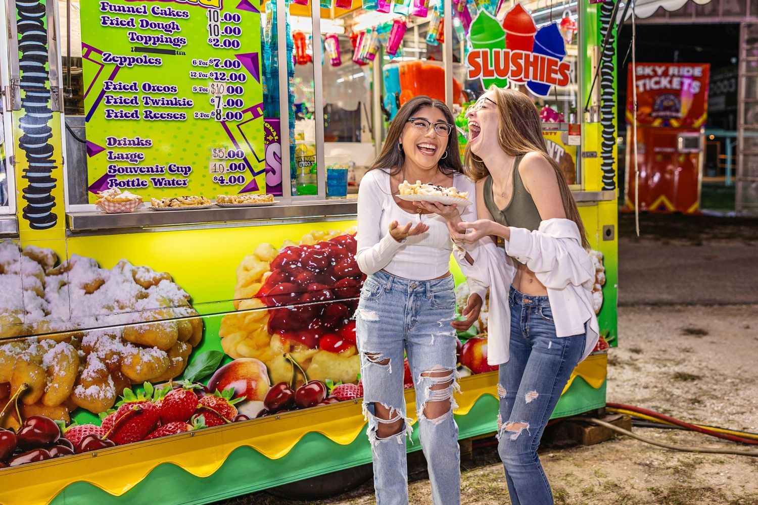 Two girls laughing and eating food at a colorful carnival fair food stand at night.