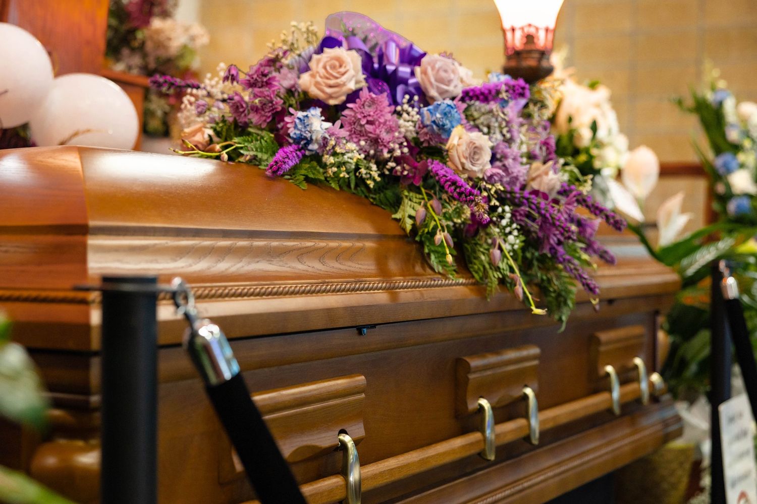 Wooden funeral casket adorned with purple and pink floral arrangement at memorial service.