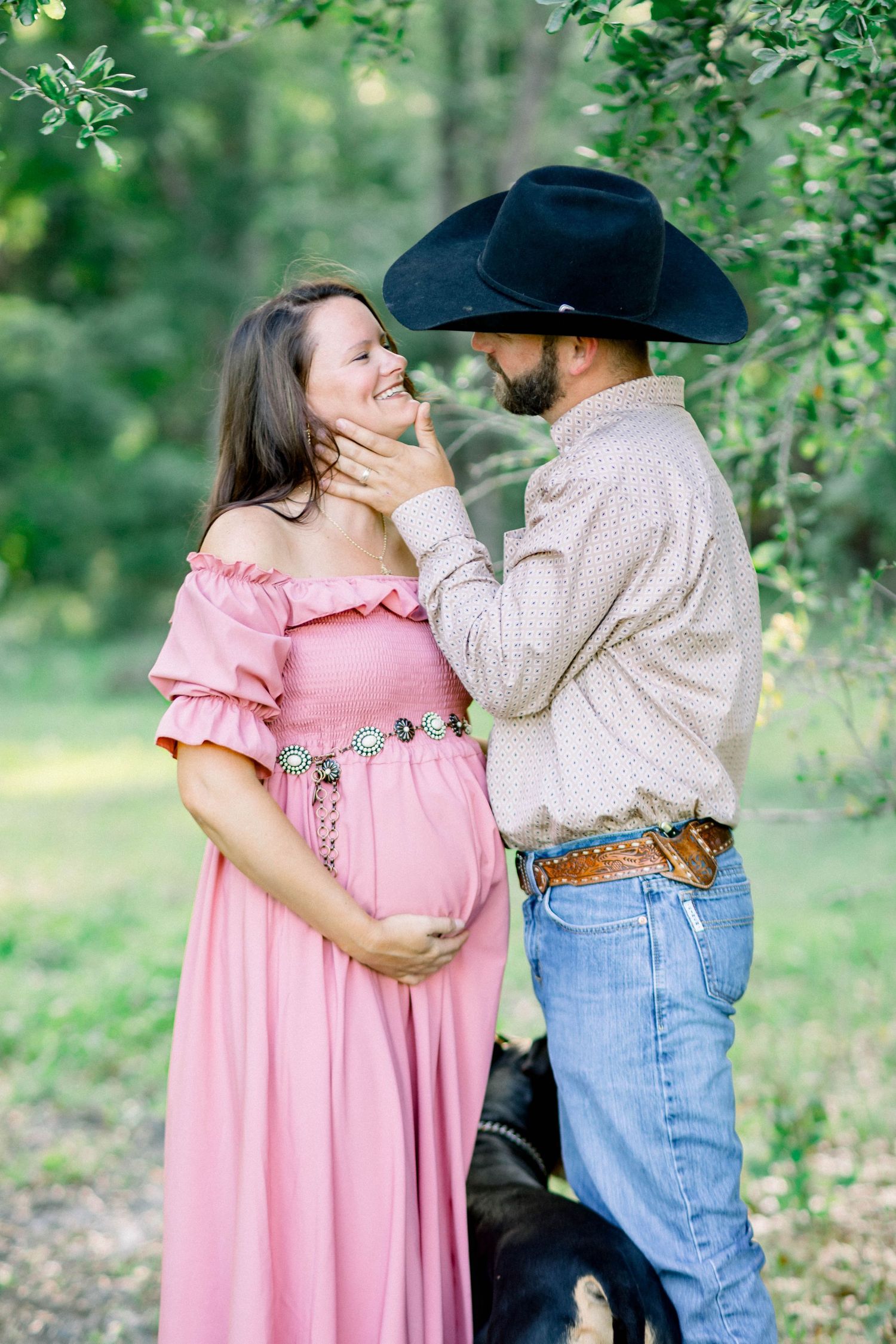 Man in black cowboy hat tenderly touching pregnant woman's face in pink dress outdoors.