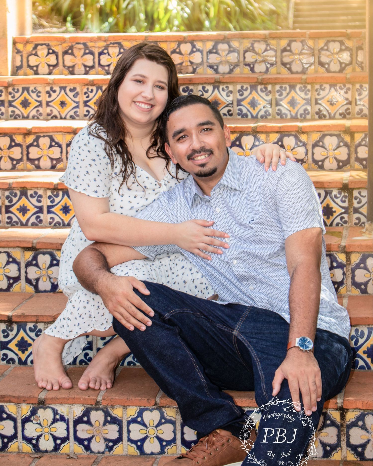 Happy couple sits on colorful Spanish tile steps with monogrammed book for engagement photos.
