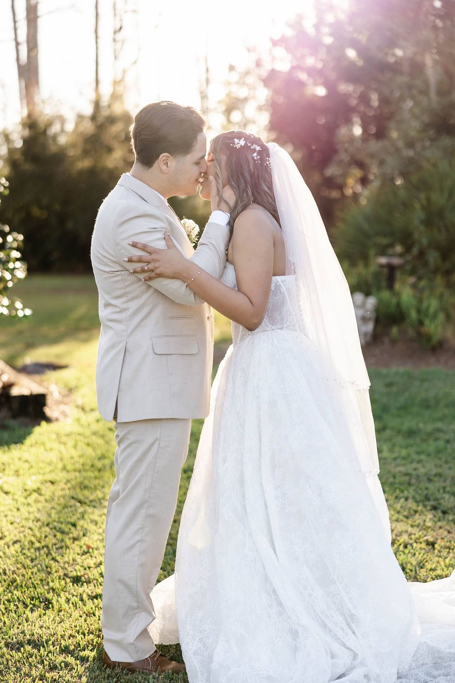 Bride in lace gown and groom in tan suit share tender moment in golden sunset garden wedding.