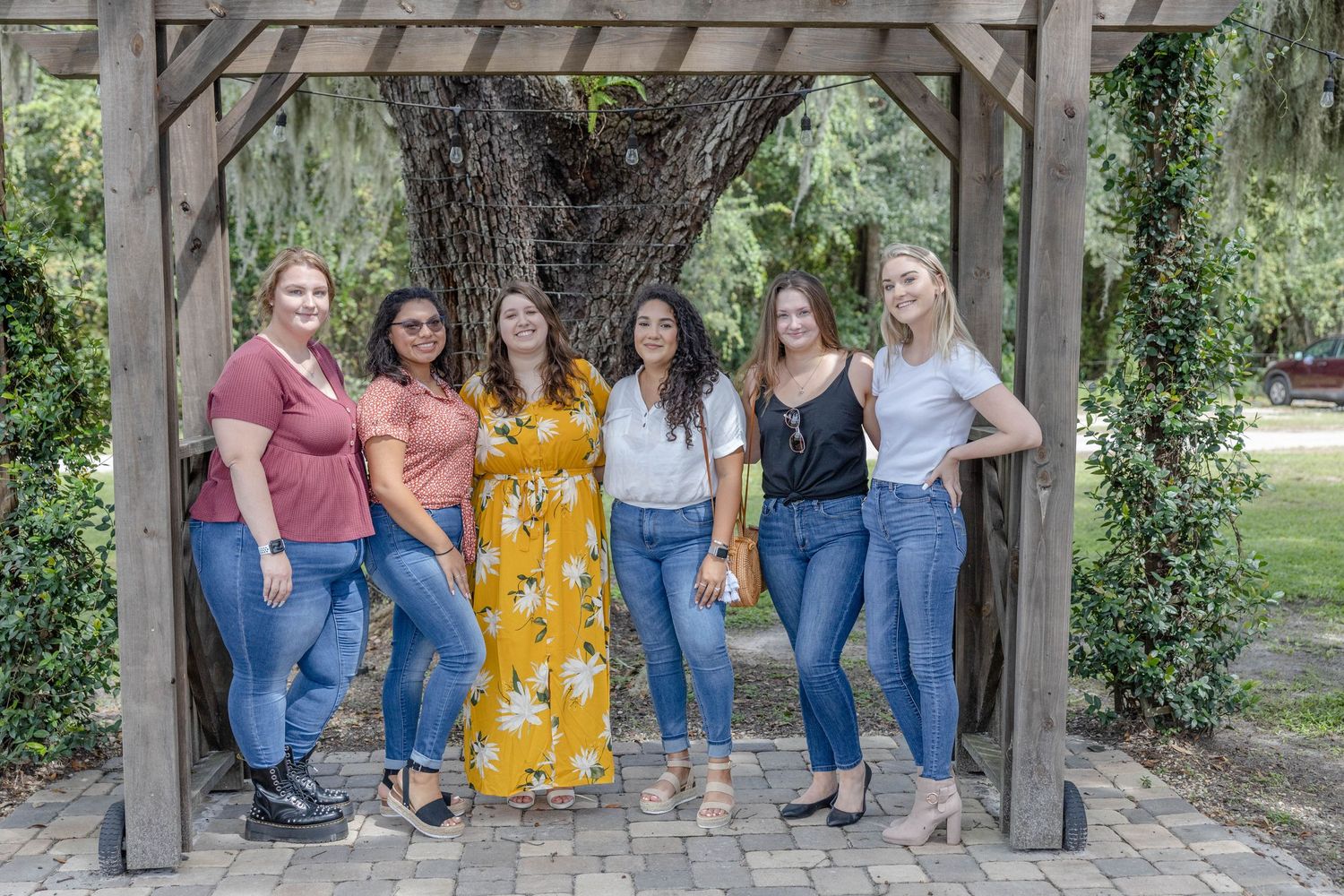 Six bridesmaids posing under wooden arbor with Spanish moss tree at outdoor wedding venue.