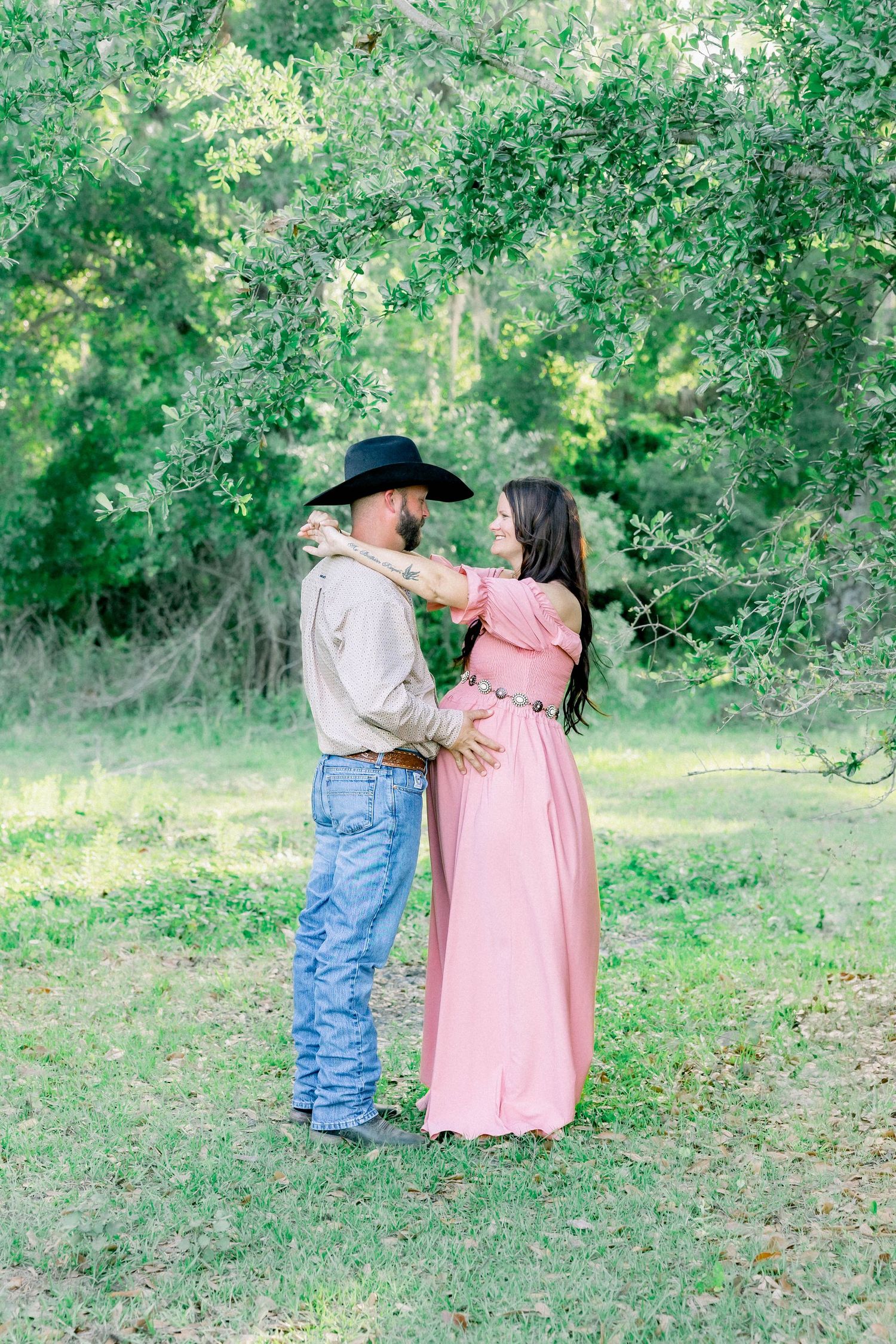 Couple dancing together in lush green field, woman in pink maternity dress and man in cowboy hat.